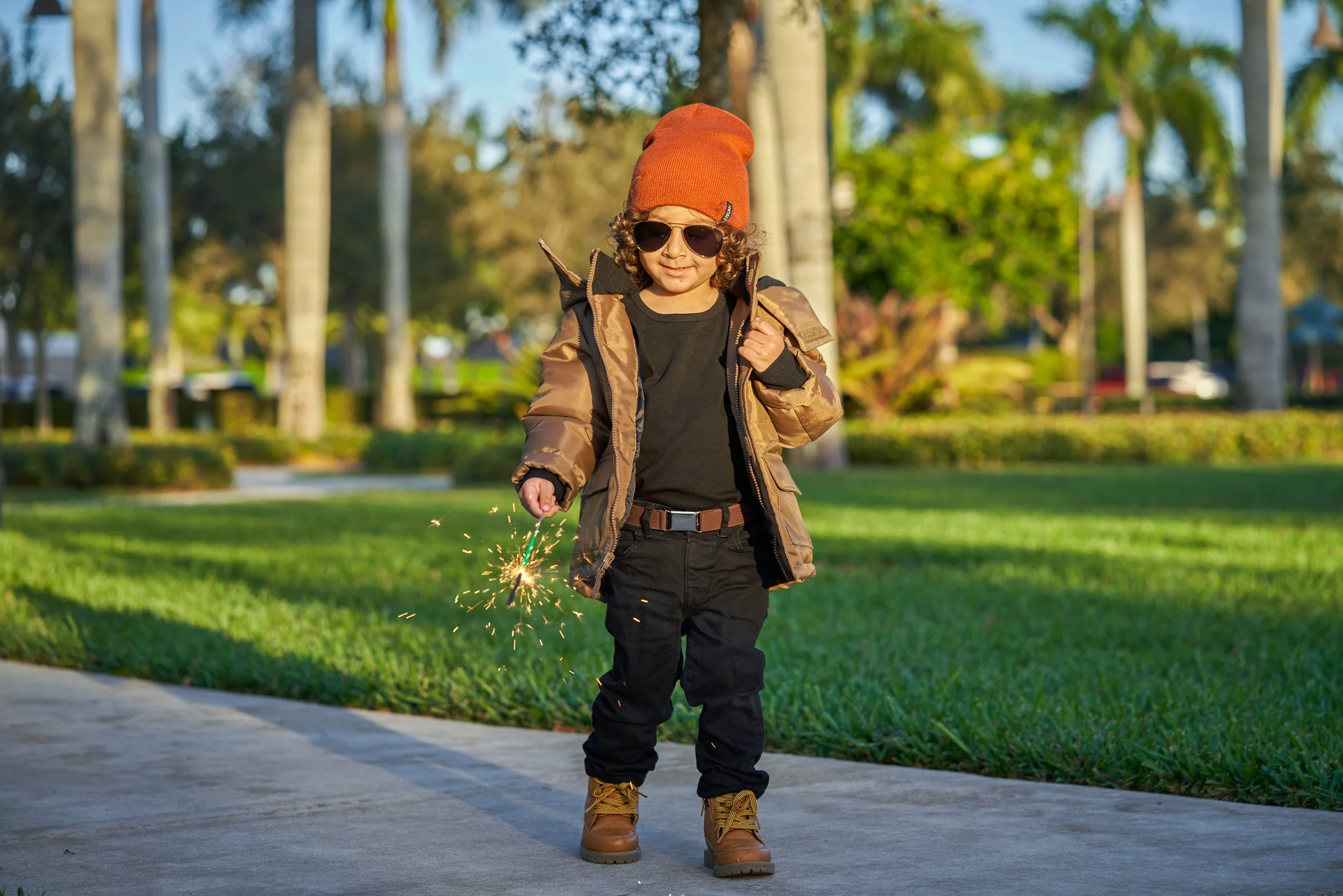 Child in sunglasses holding a sparkler, wearing a brown jacket, black clothing, brown boots and an orange hat, walking on a sidewalk in a park with green grass and trees.