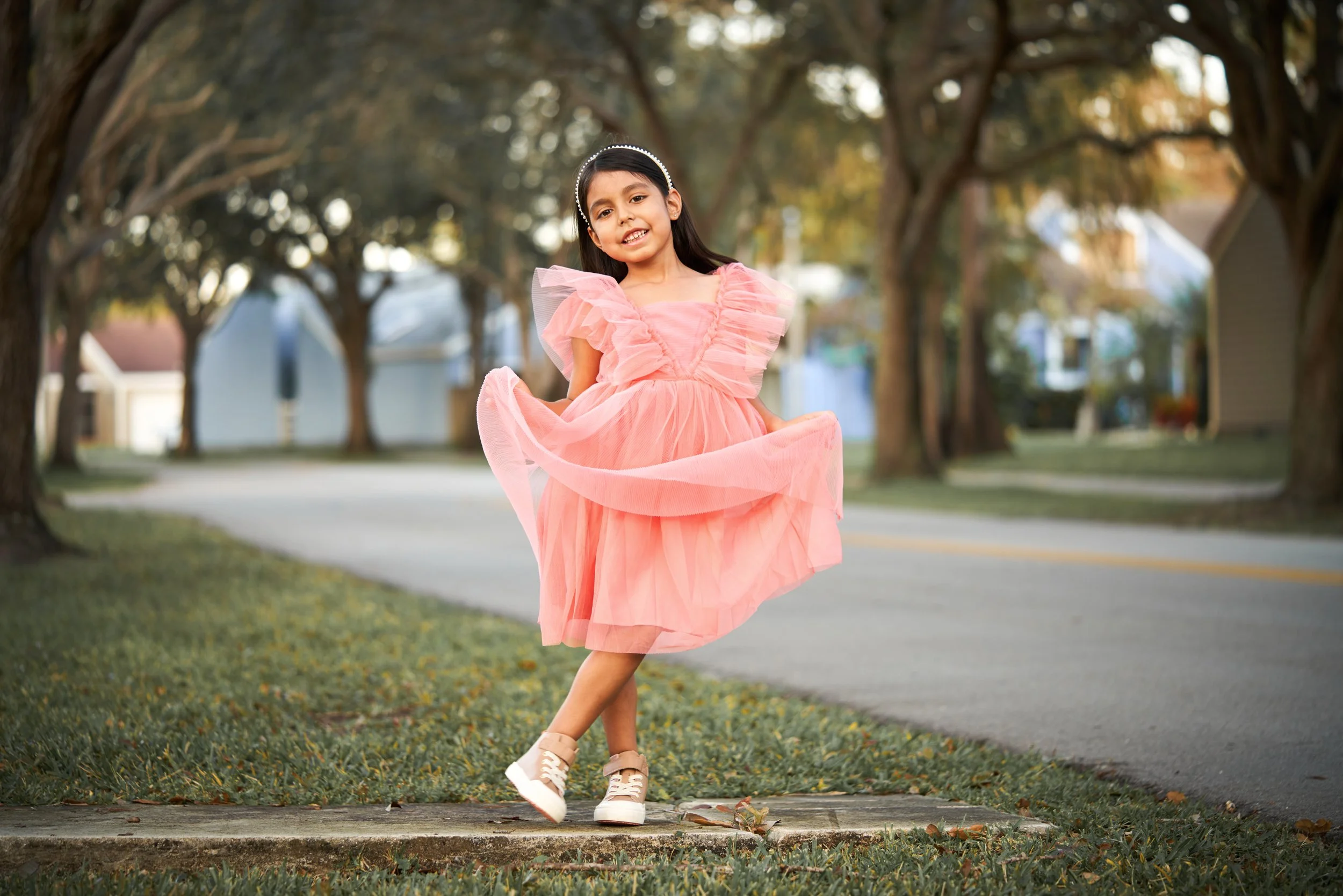 A young girl in a pink dress standing on a sidewalk, lifting her dress and smiling outdoors in a neighborhood with trees and houses.