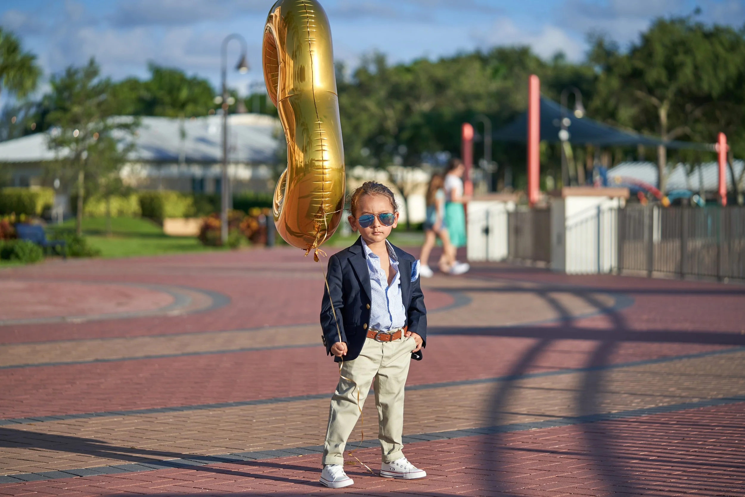 Young boy in a navy blazer and khakis holding a gold balloon, standing on a brick pathway in a park, wearing sunglasses, with other park visitors in the background.
