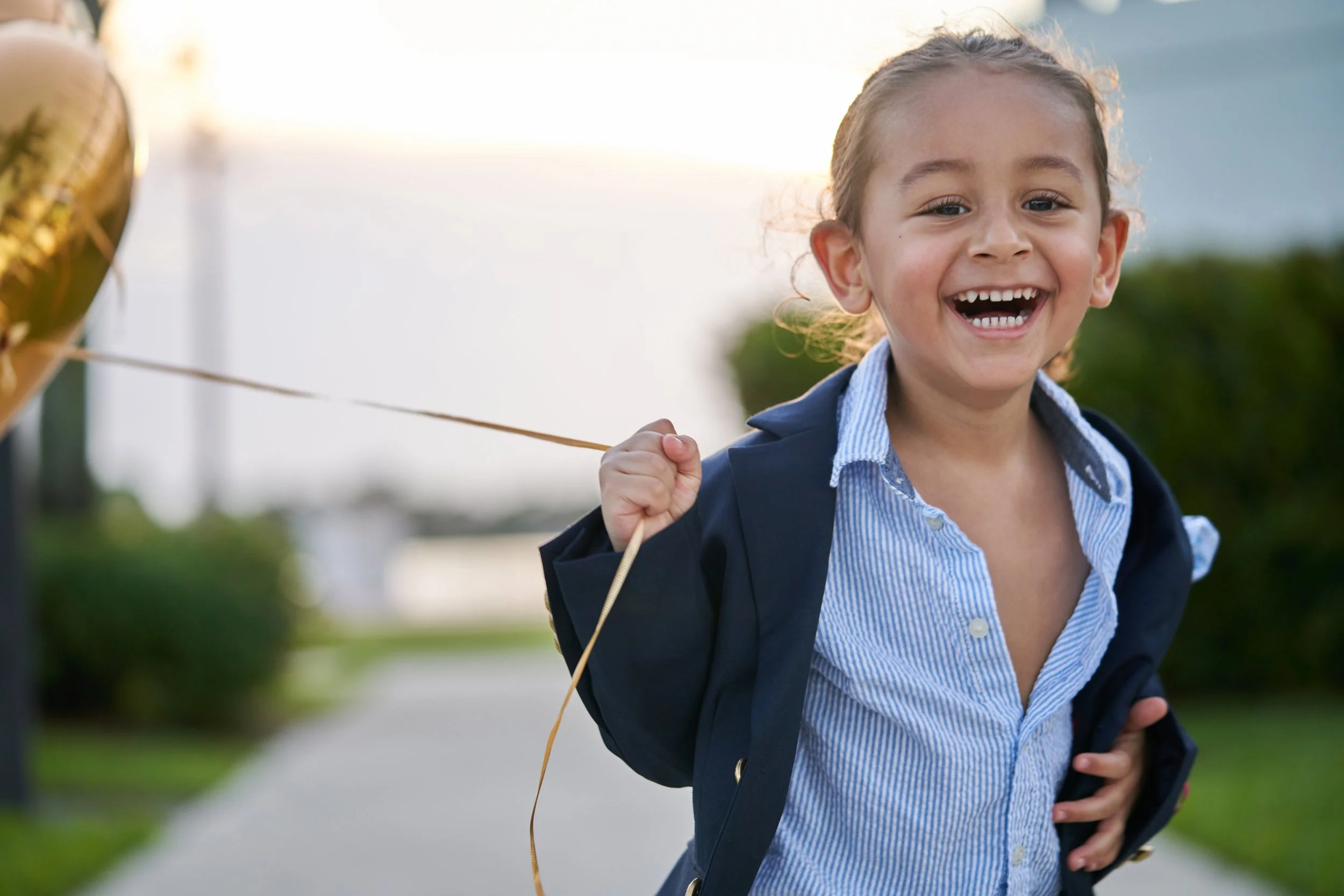 A young girl with curly hair, smiling and laughing, holding a gold balloon string, outdoors on a sidewalk with greenery.