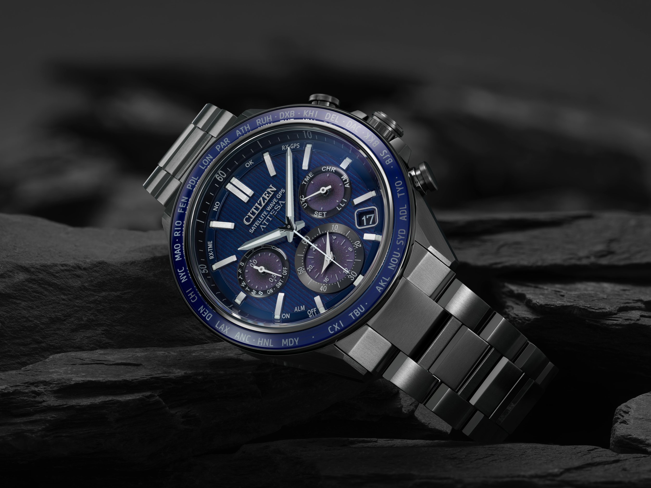 A Citizen satellite watch with a blue dial, silver case, and metal band resting on dark rocks.