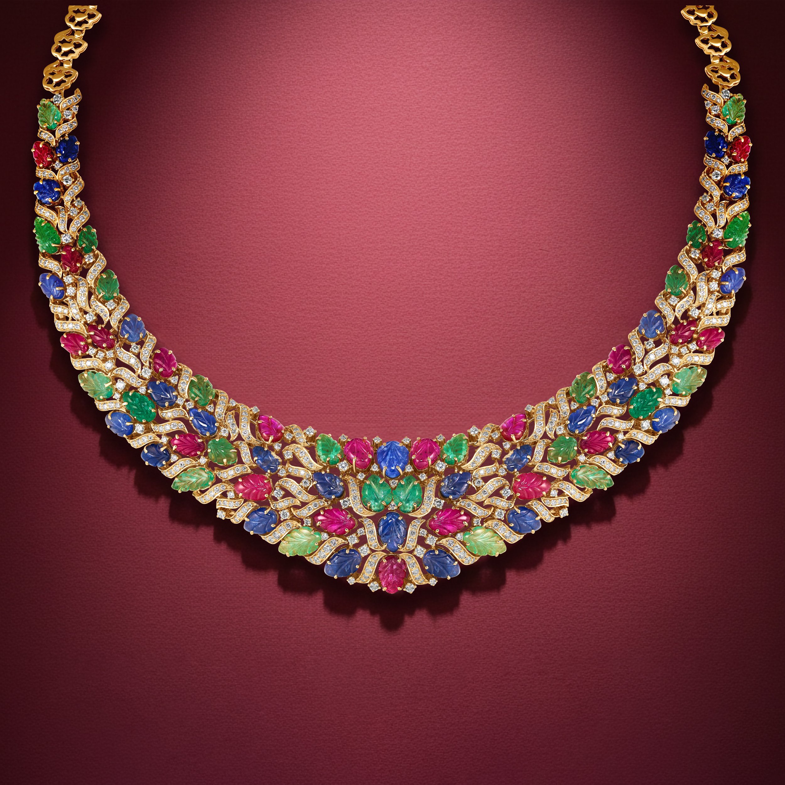 A colorful diamond necklace with green, red, blue, and pink gemstones, set in gold, displayed against a maroon background.