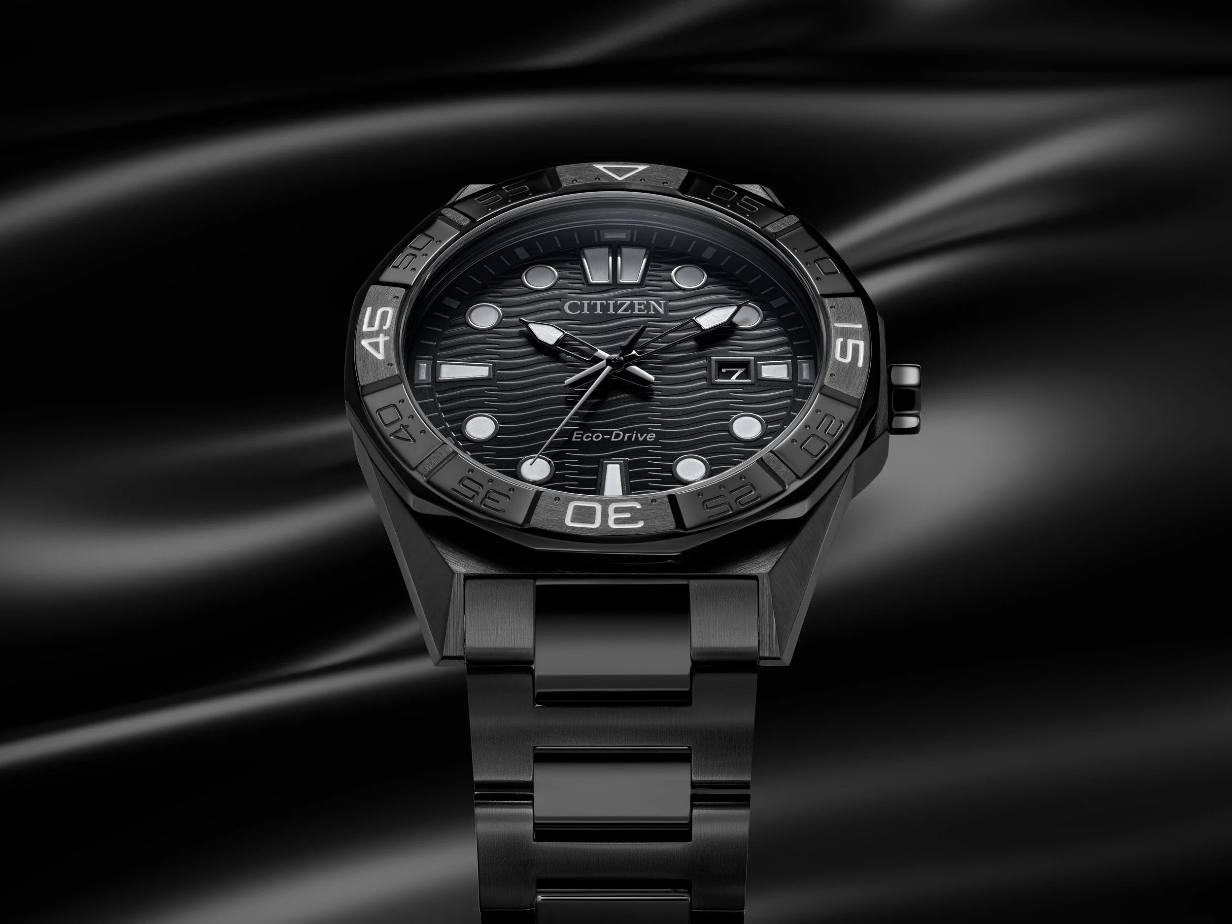 A black Citizen Eco-Drive wristwatch with a black band and textured black dial, set against a black satin background.