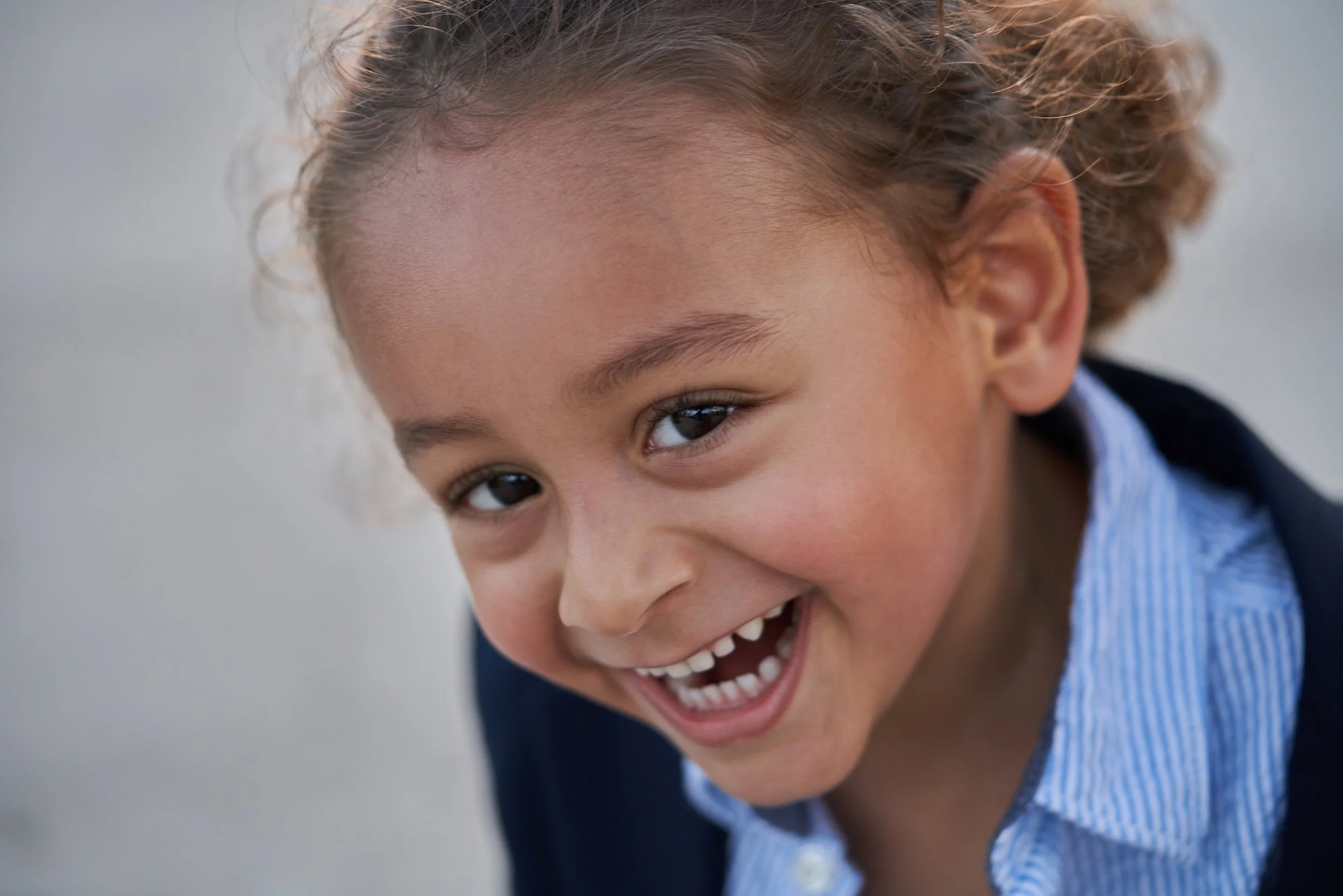 A young child with curly hair smiling happily, showing teeth, wearing a blue shirt and a dark jacket.