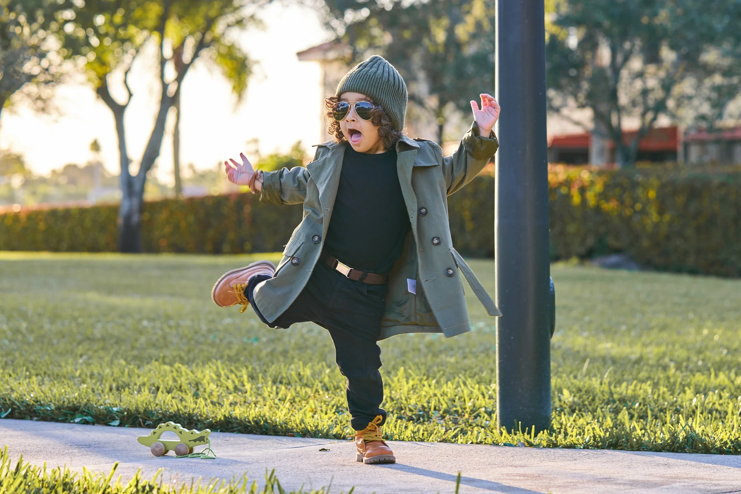 Young child wearing sunglasses, a beanie, and a coat, balancing on one leg in a park during sunset with a toy car on the sidewalk.
