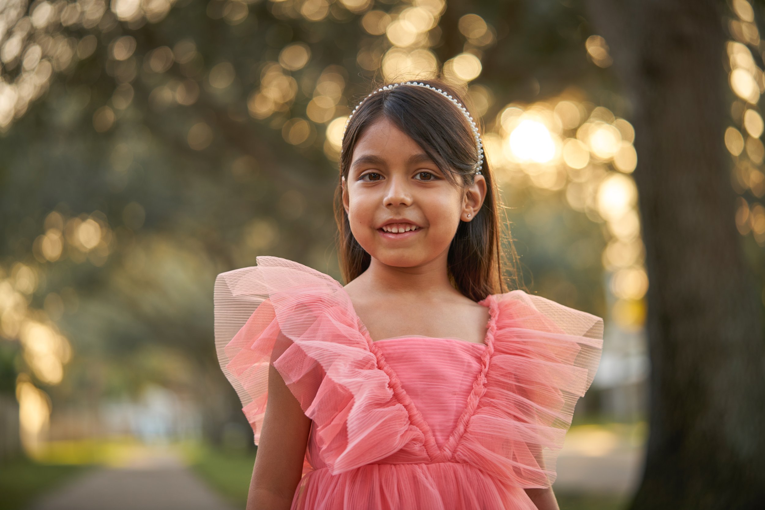 A young girl wearing a pink dress with ruffled tulle sleeves, standing outdoors with trees and blurred sunlight in the background.
