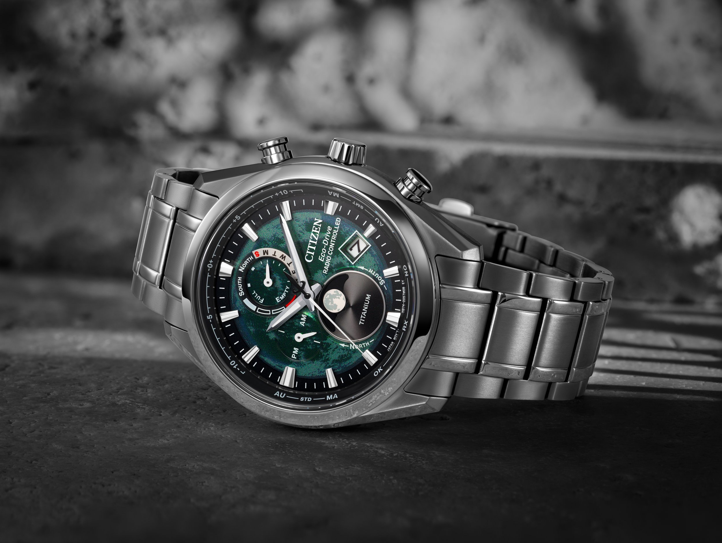 A Citizen Eco-Drive radio-controlled titanium wristwatch with a metal band, placed on a dark textured surface in front of a blurry stone background.