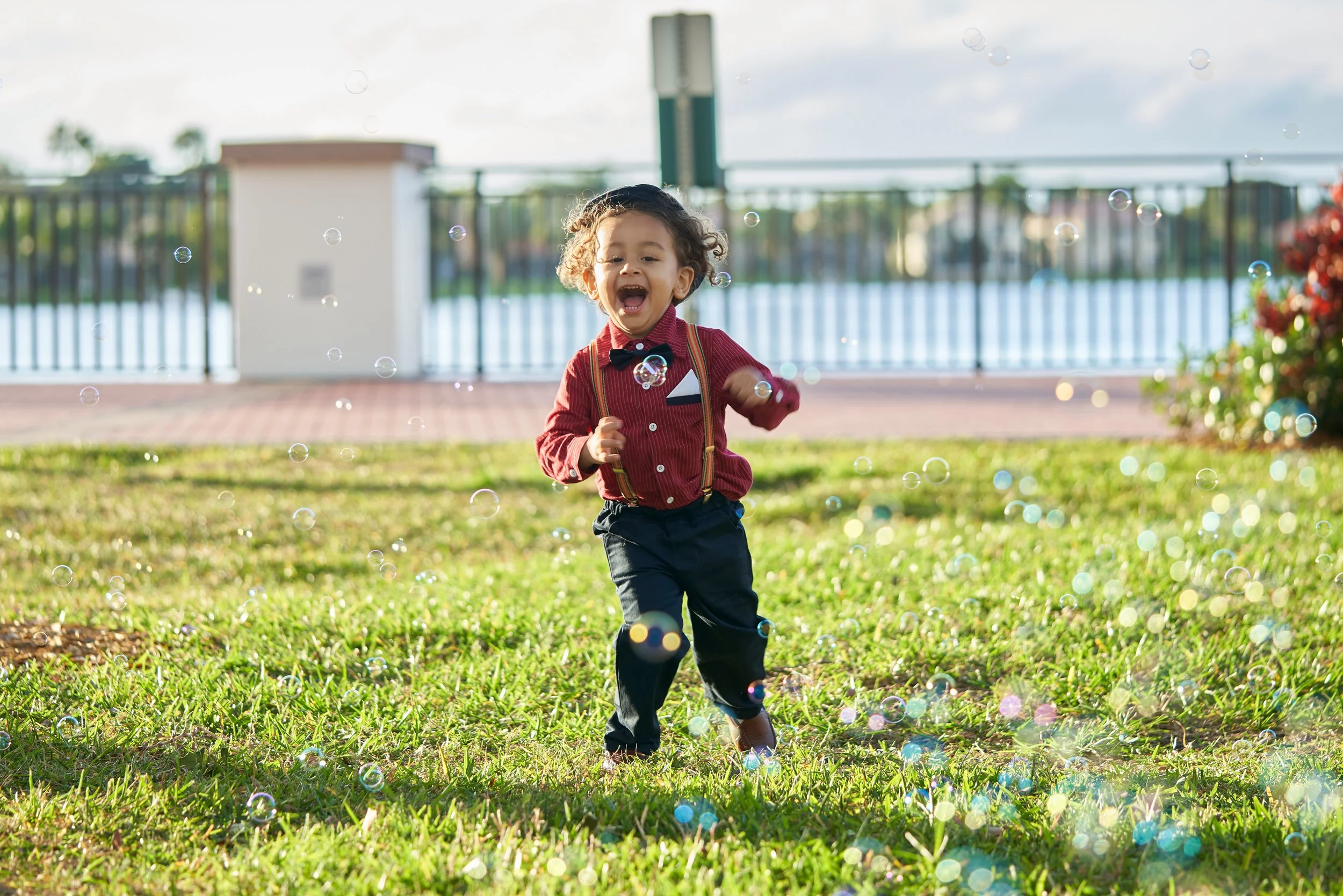 A young boy with curly hair running through a grassy area, smiling and playing among soap bubbles, near a waterfront with a railing and a small building in the background.