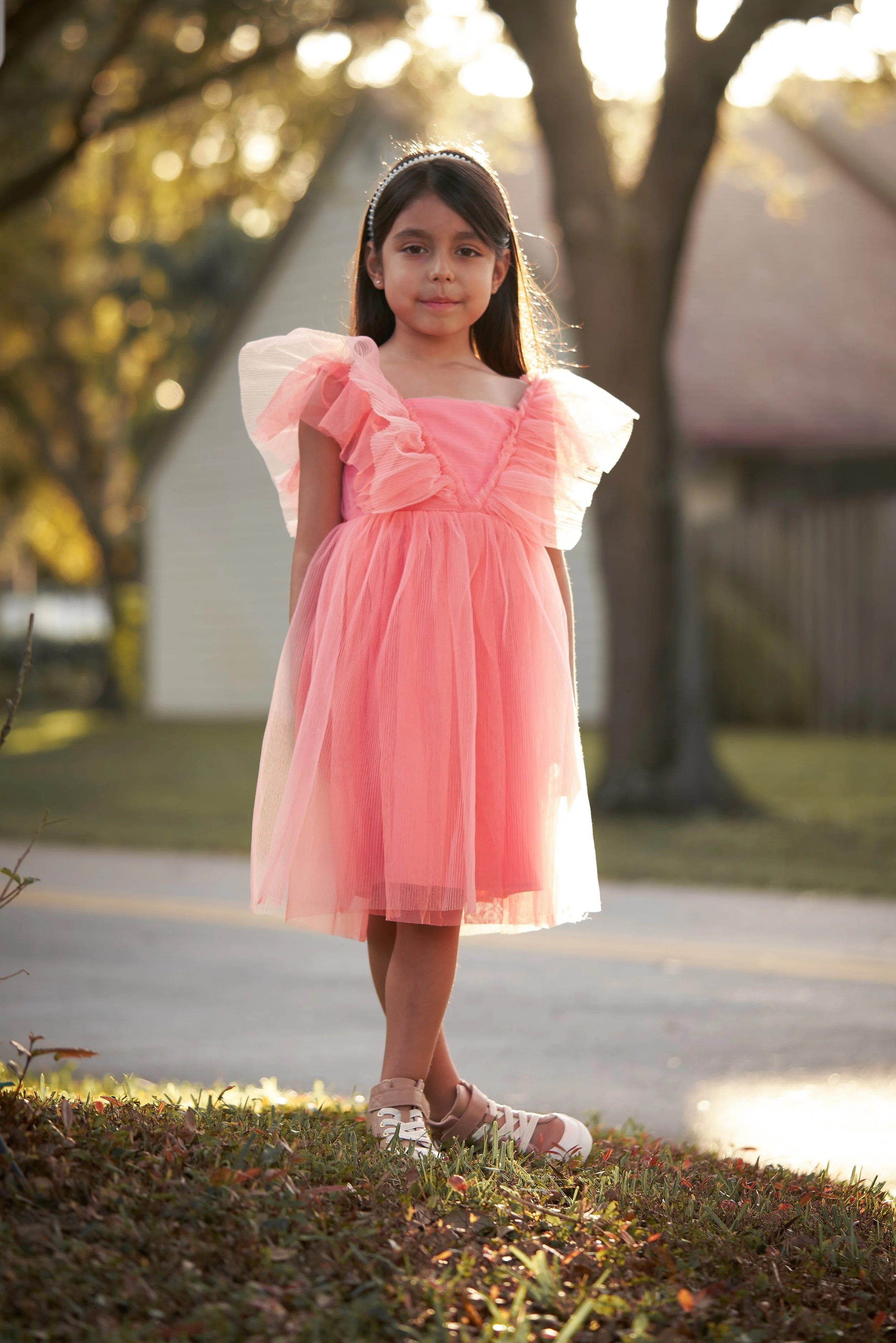 A young girl in a pink dress standing outdoors in the late afternoon with trees and houses in the background.
