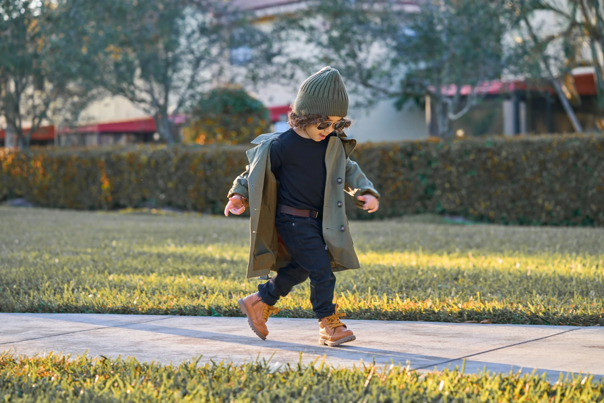 A young boy with curly hair, wearing sunglasses, a green beanie, black shirt, green jacket, black pants, and tan boots, is skating or playing on a sidewalk in a park or yard with grass, bushes, and trees in the background during daytime.