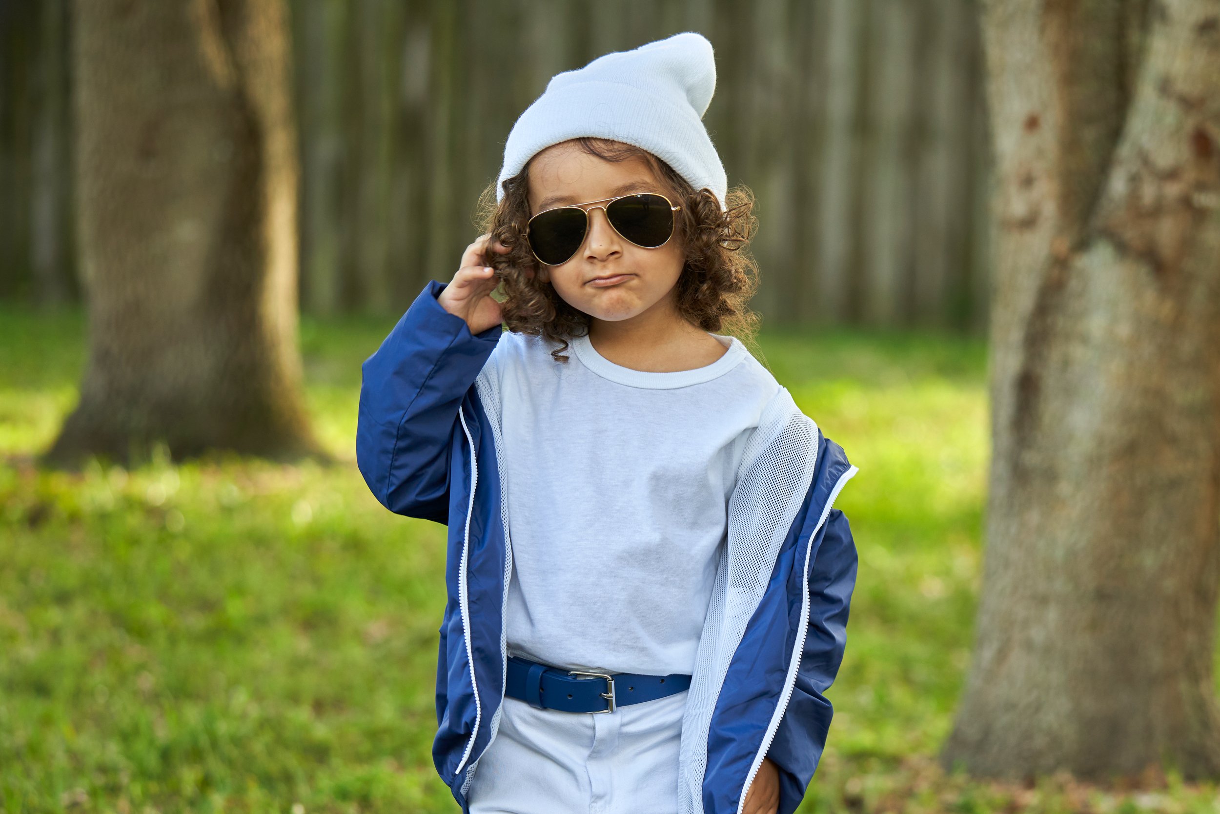A young child with curly hair wearing sunglasses, a light blue beanie, a blue jacket, and white pants standing outdoors in a wooded area.