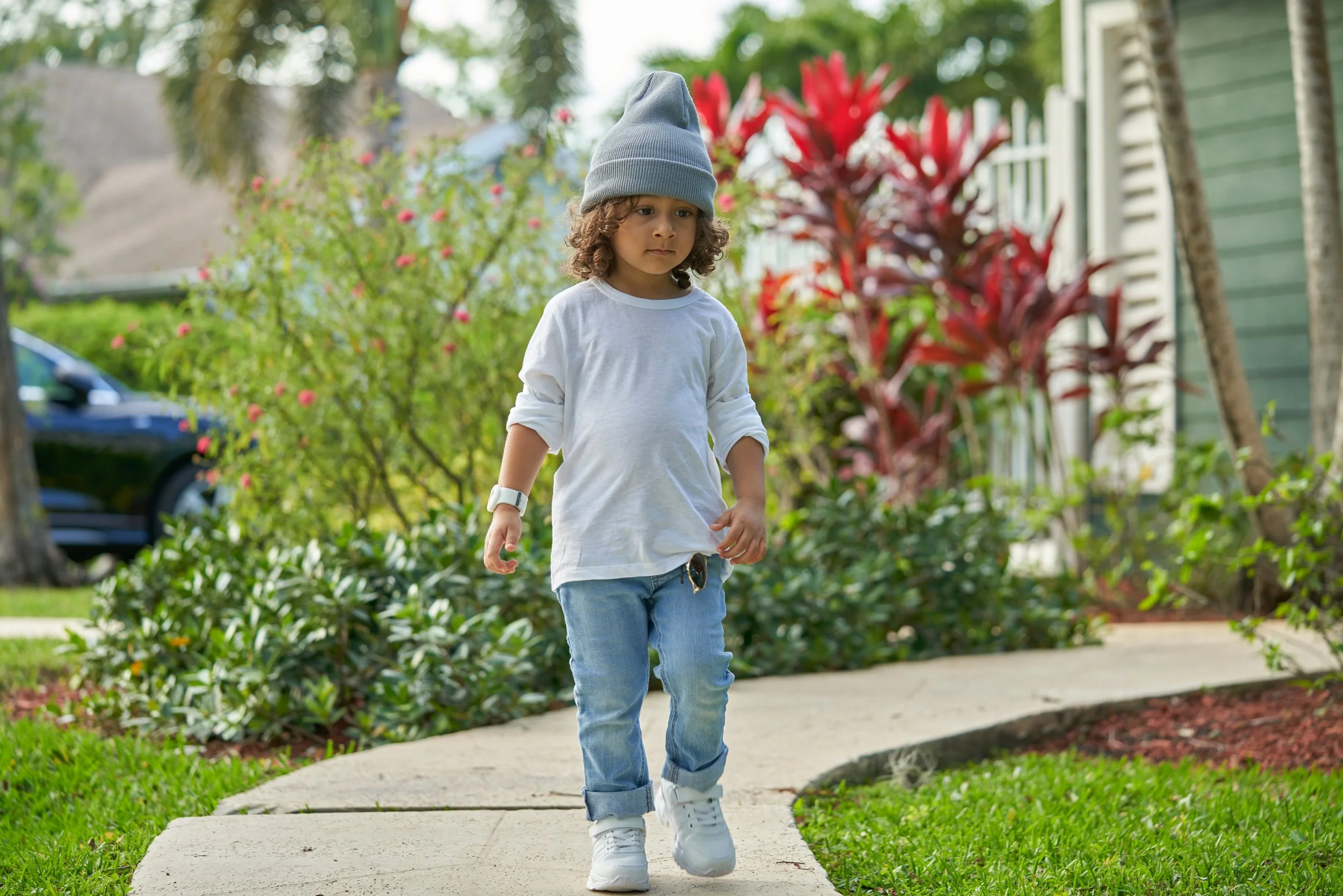 Young child walking on a sidewalk in a green yard with plants and trees, wearing a gray beanie, white long sleeve shirt, jeans, and sneakers.