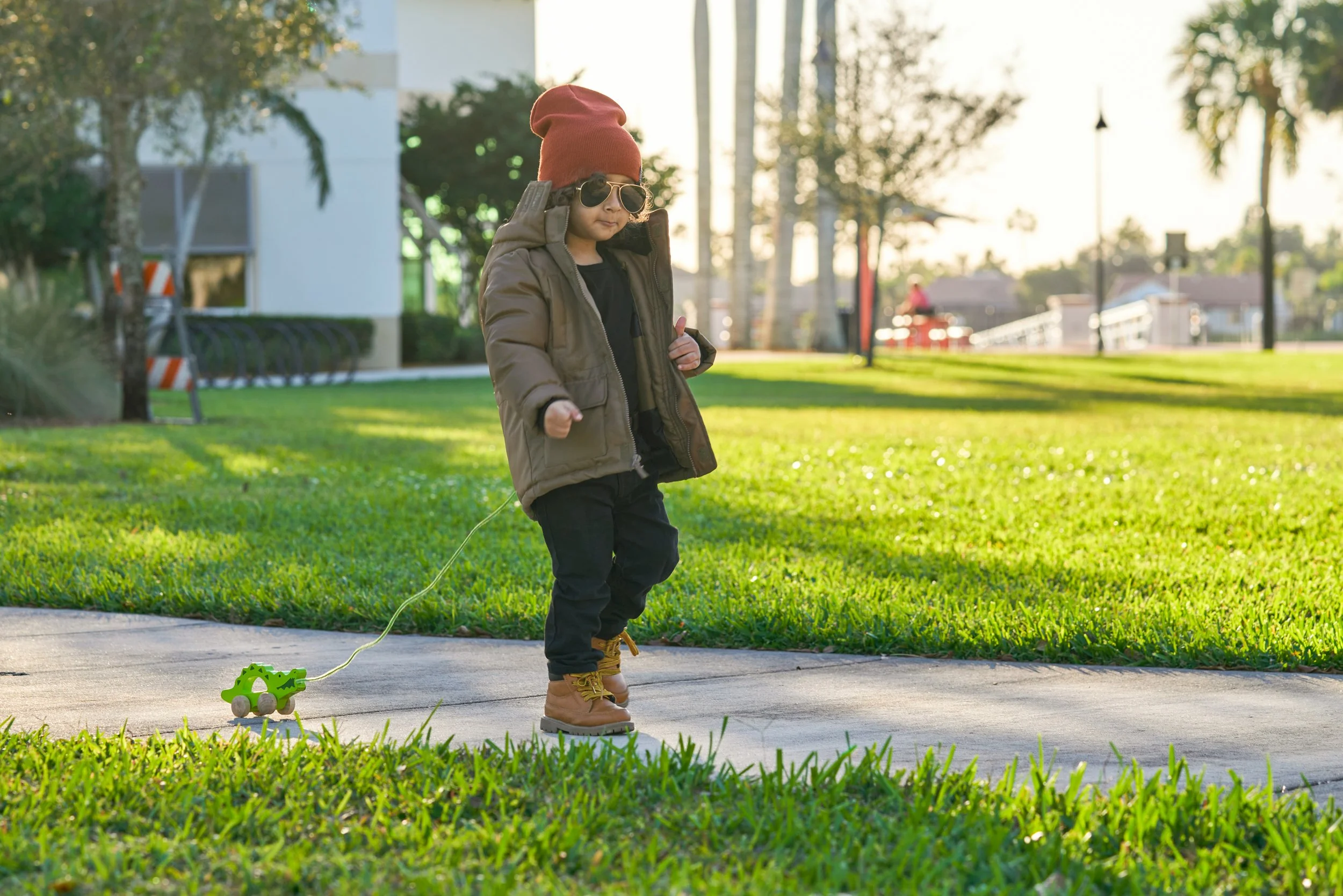 A young boy wearing sunglasses, a red beanie, a brown jacket, black pants, and tan boots is walking on a sidewalk pulling a green toy figure on wheels with a string, in a park during late afternoon.