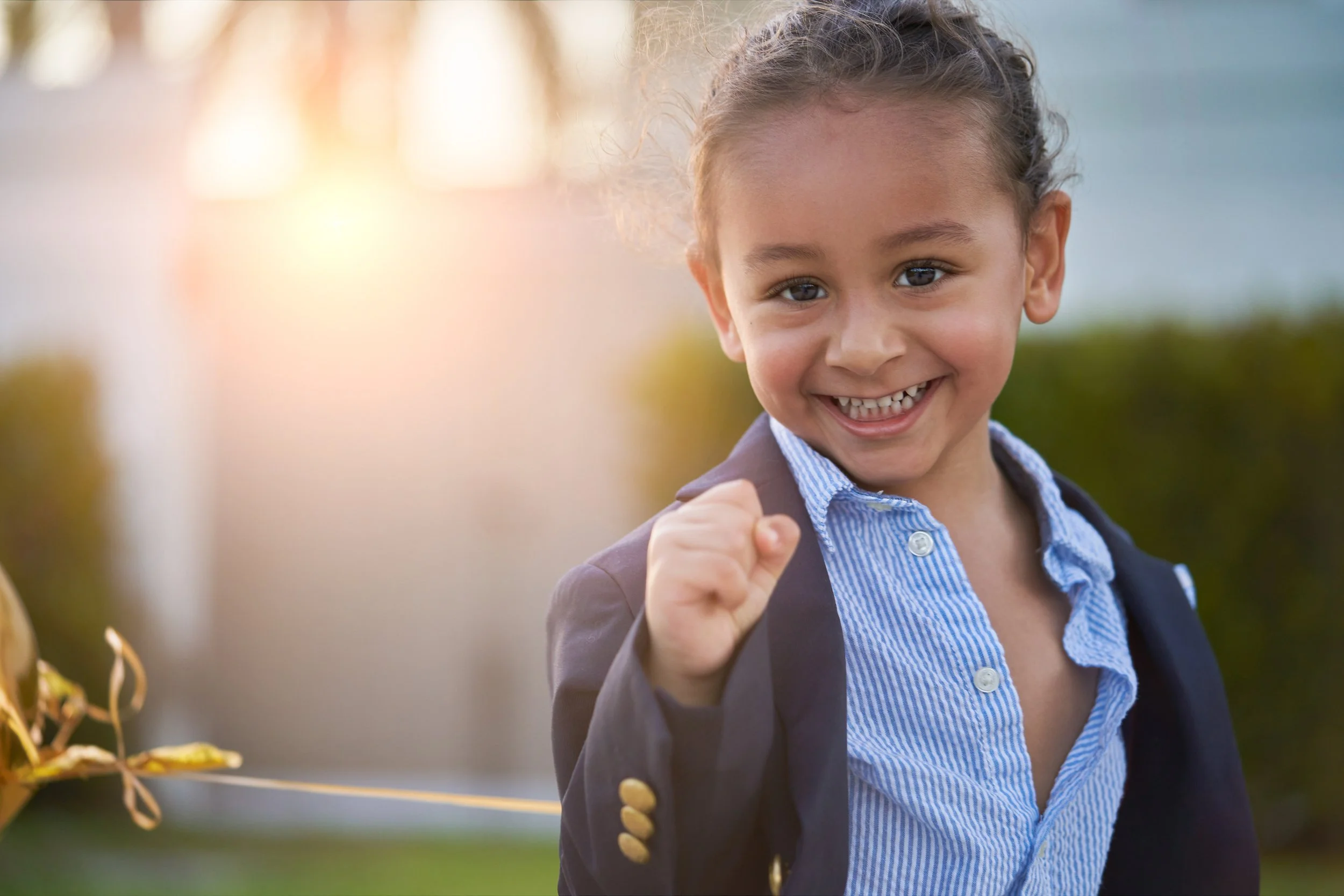 A young girl with curly hair, smiling and showing a fist pump gesture, outside during sunset.