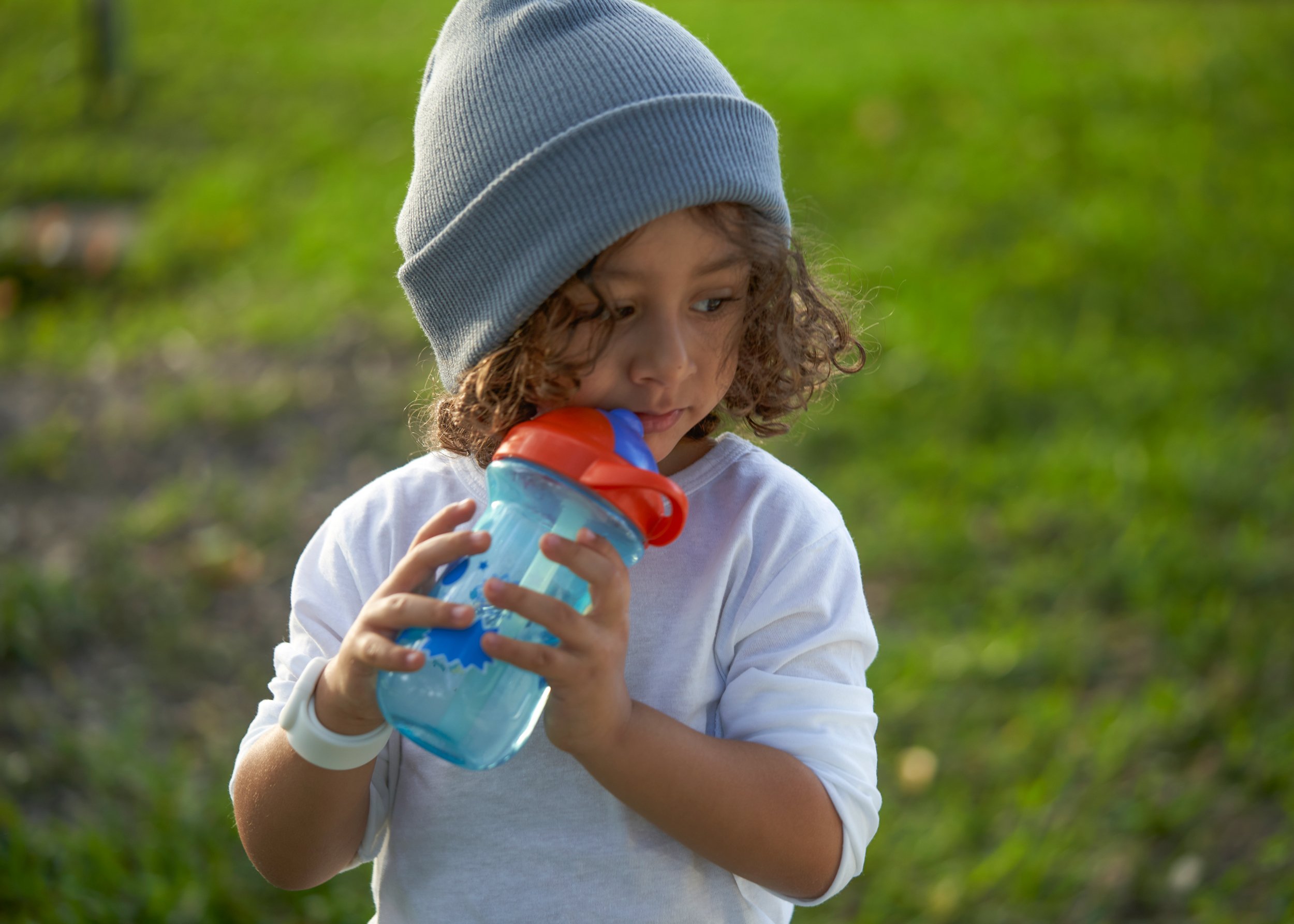 Young boy with curly hair wearing a gray beanie and white shirt holding a sippy cup outdoors.