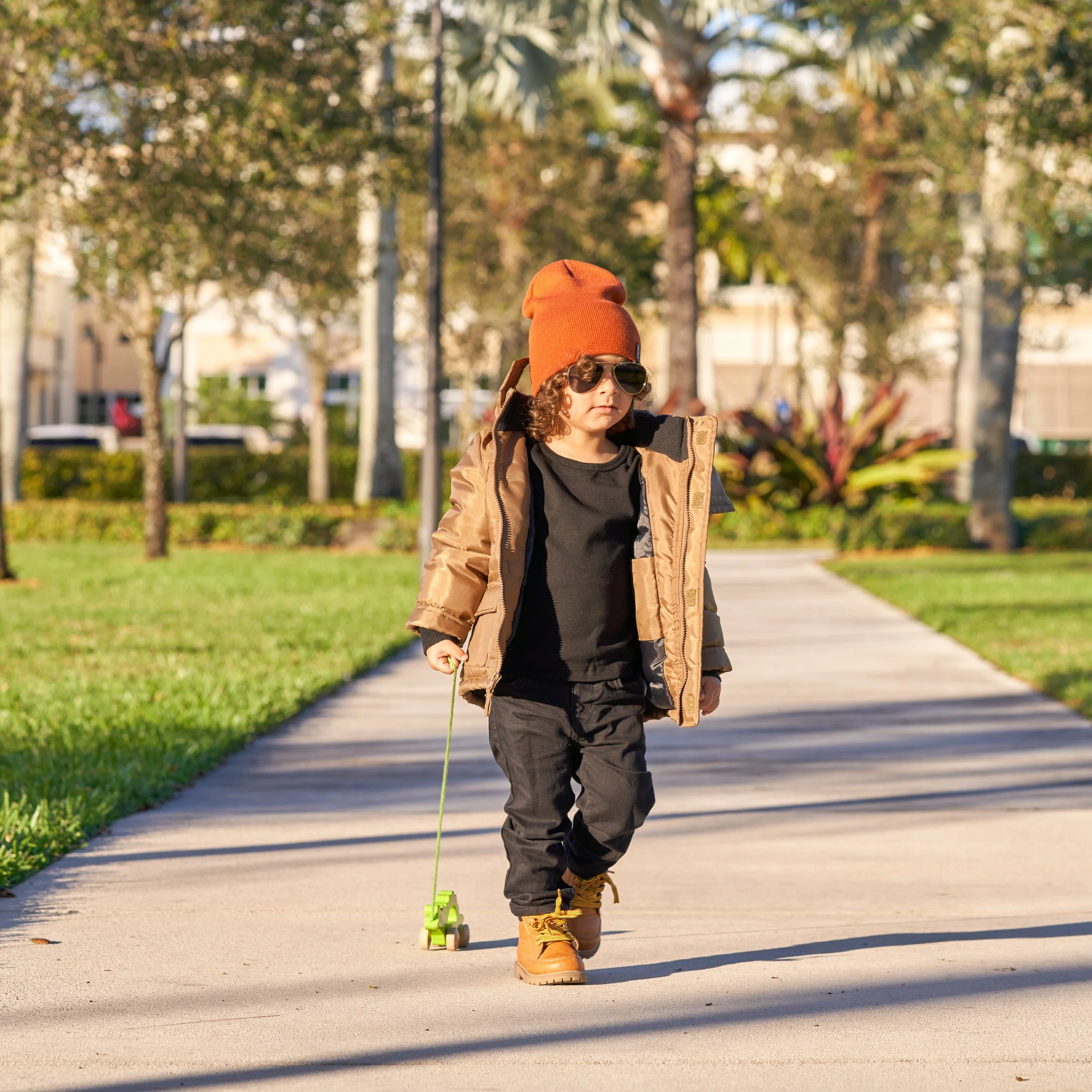 Young boy walking on a sidewalk wearing sunglasses, a brown jacket, a black shirt, pants, a red beanie, and boots, pulling a small green toy car with a string, in a park with trees and buildings in the background.