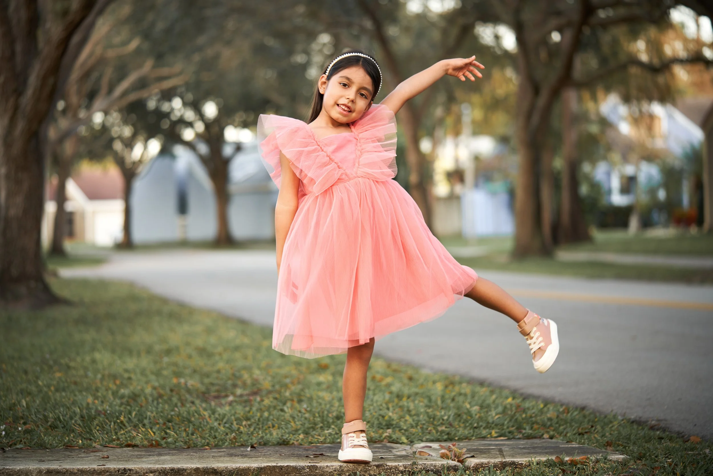 A young girl in a pink dress and sneakers posing playfully on a sidewalk outdoors, with trees and houses in the background.