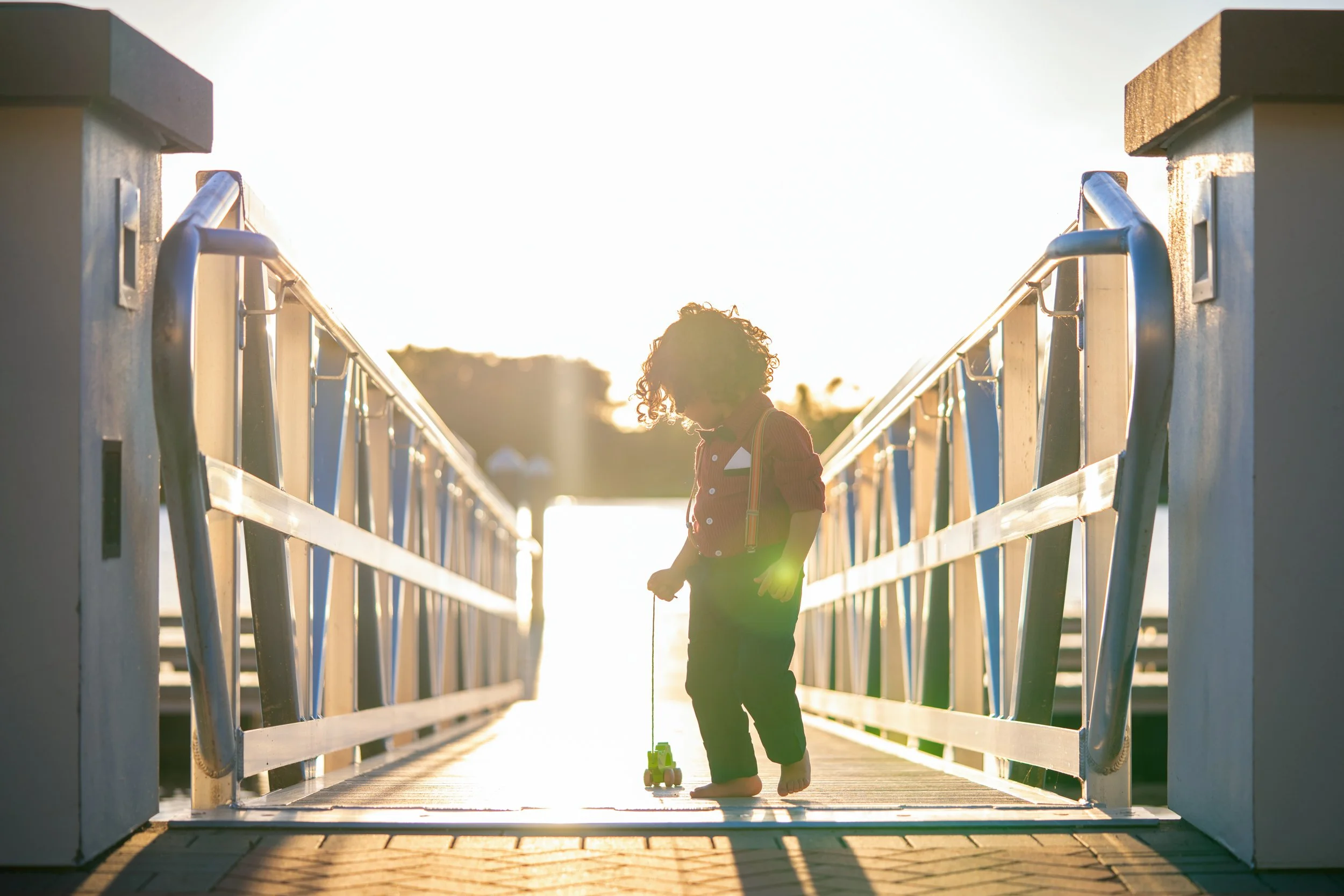 Child with curly hair playing golf on a dock at sunset, wearing suspenders and standing barefoot.