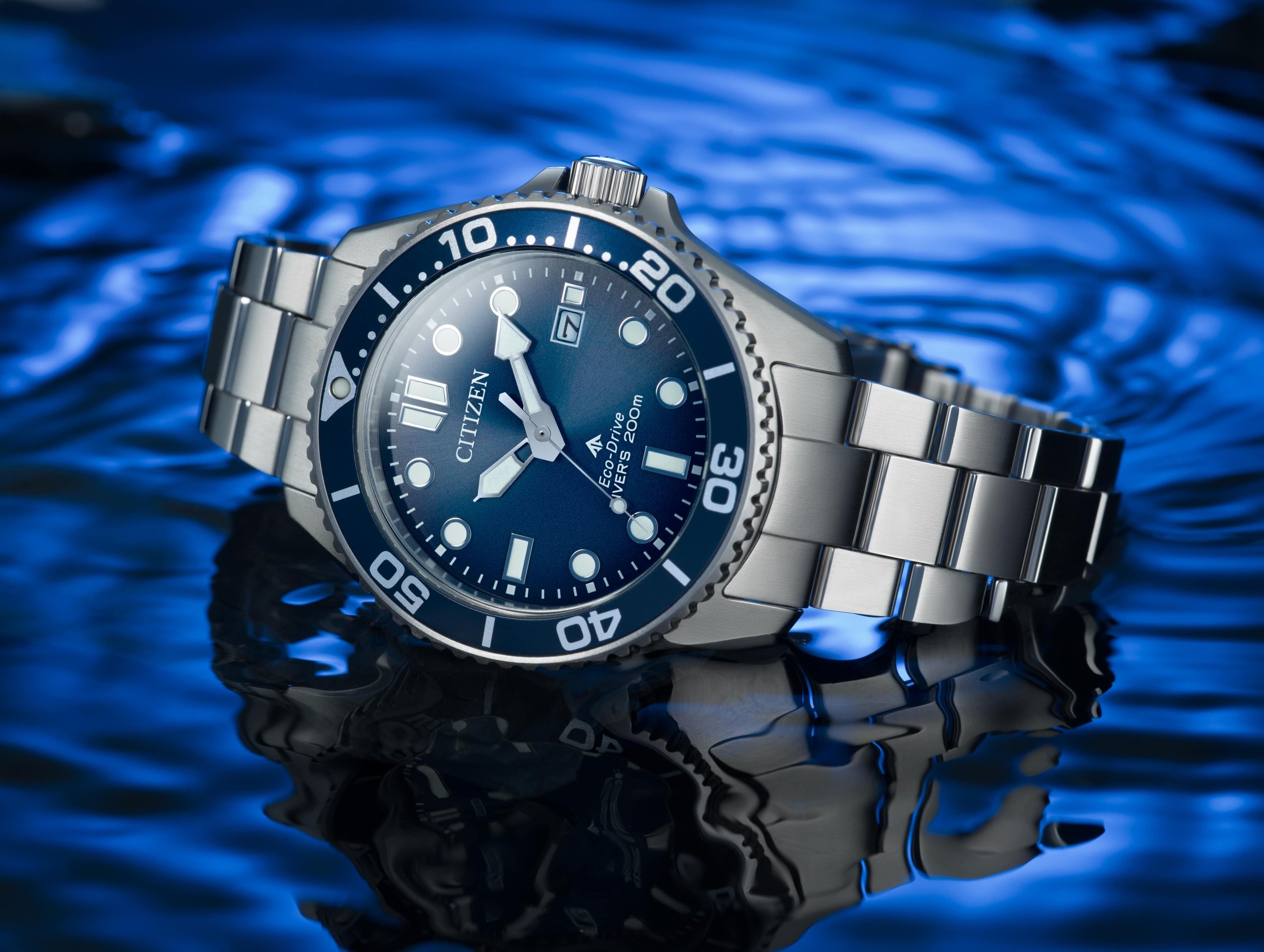A Citizen Eco-Drive diver's watch with a blue face and bezel, silver band, resting on a water surface with reflections.