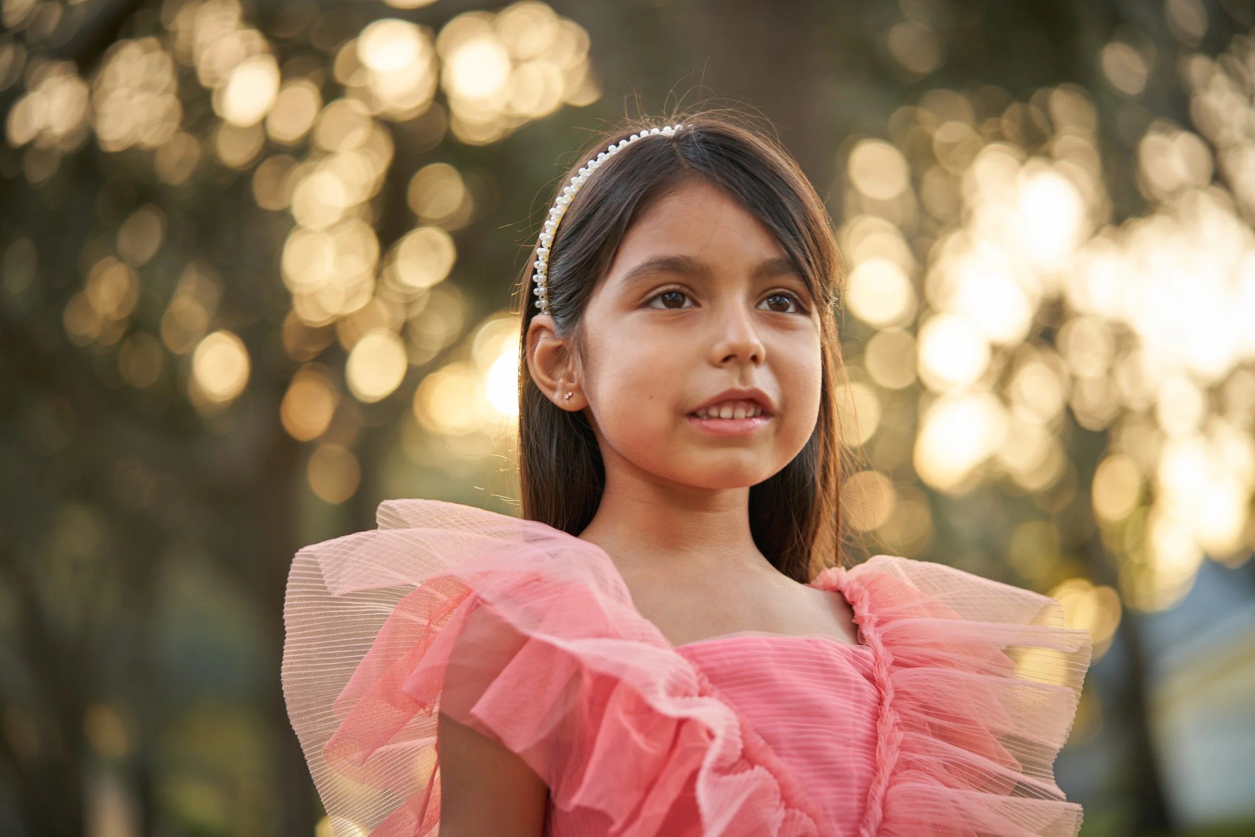 Young girl wearing a pink ruffled dress and a pearl headband, standing outdoors with bokeh sunlight behind her.
