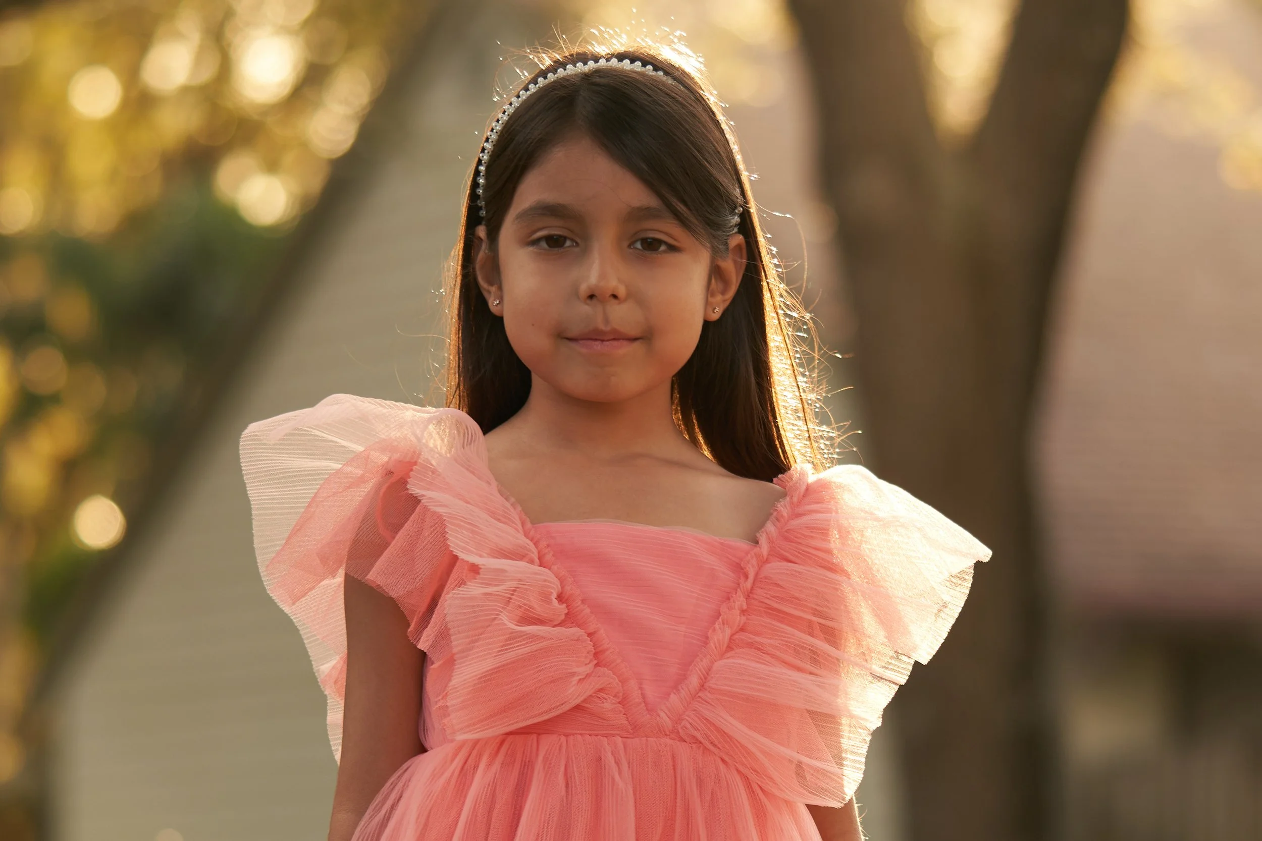 A young girl in a pink ruffled dress standing outdoors with trees in the background, illuminated by warm sunlight.