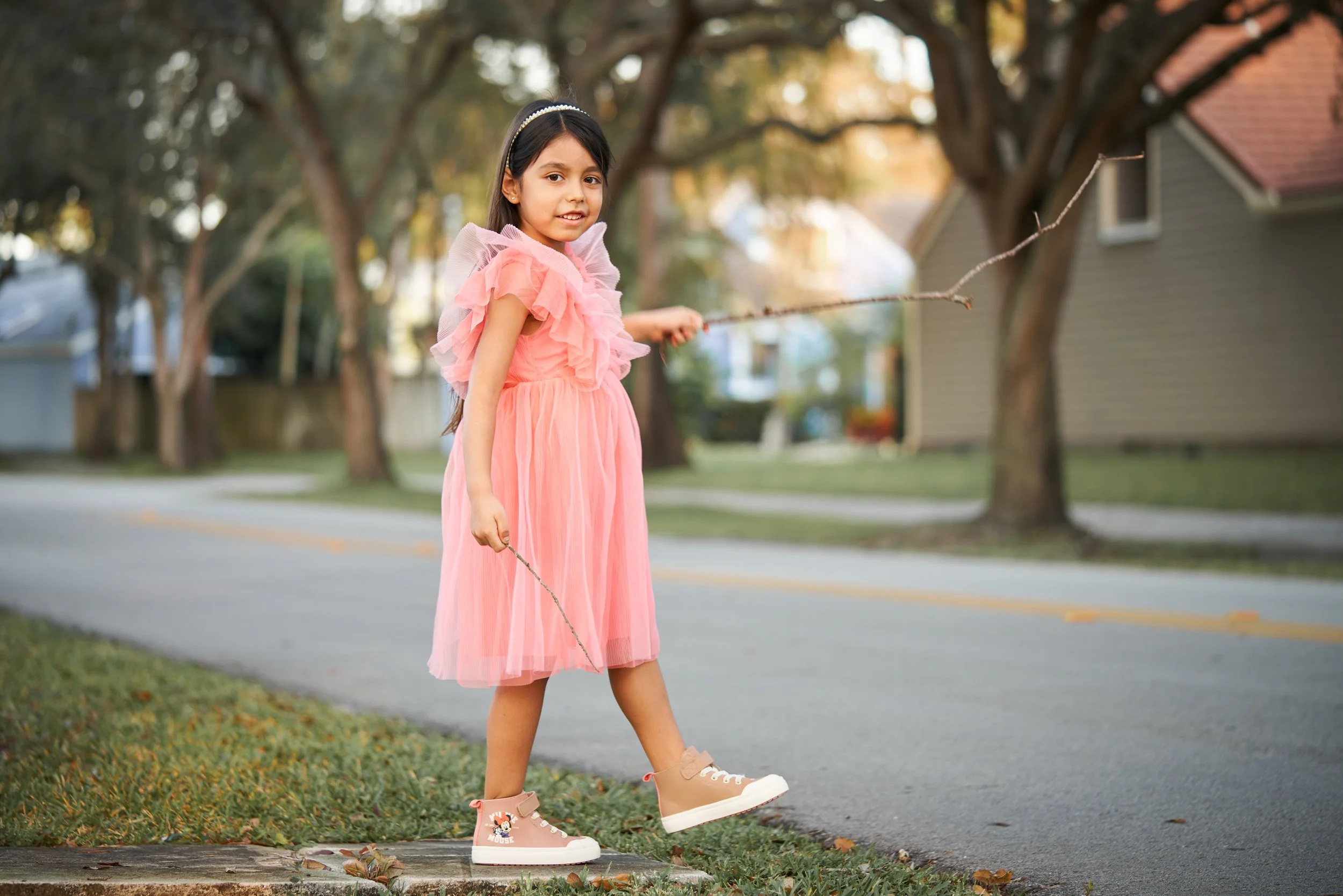 A young girl in a pink dress and pink sneakers standing on the sidewalk holding sides of a small stick, outdoors with trees and houses in the background.