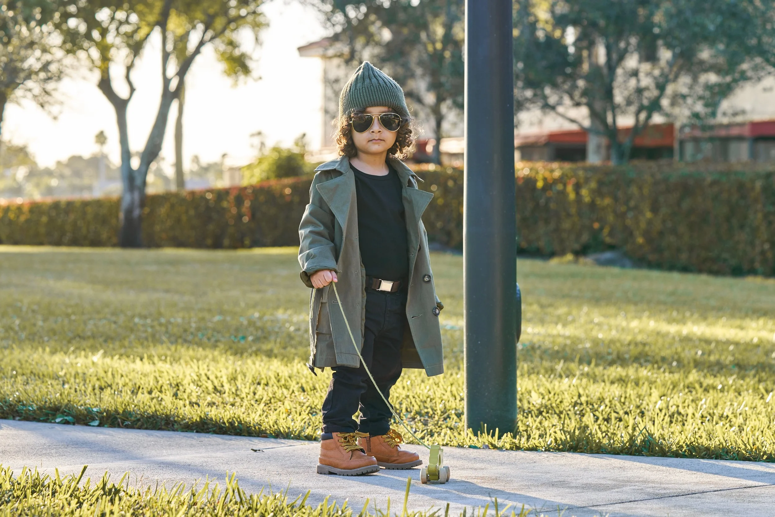 Young boy with curly hair wearing a beanie, sunglasses, a trench coat, and boots, standing on a sidewalk holding a scooter.