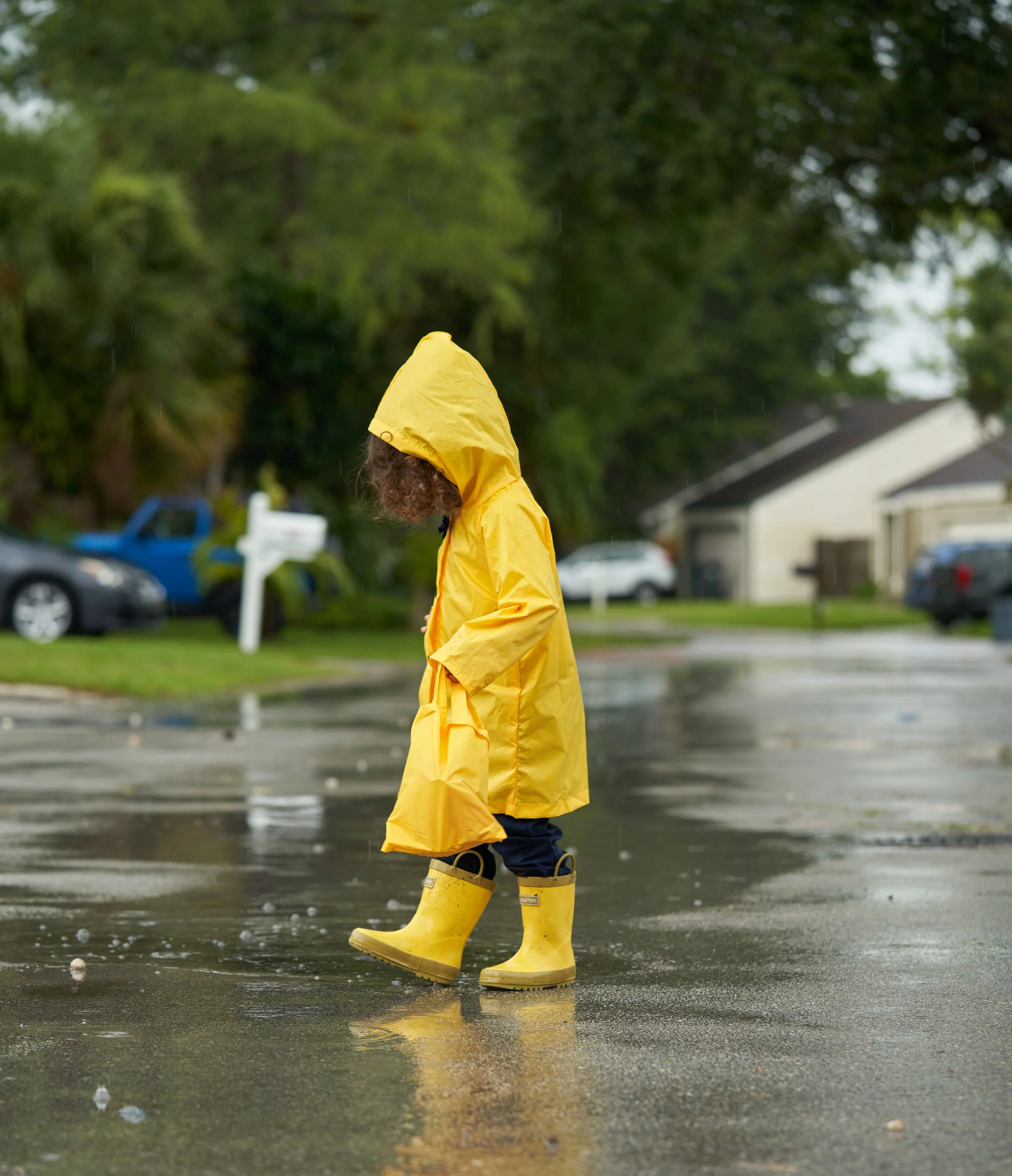 Child in yellow raincoat and rain boots walking on wet street during rainy day.
