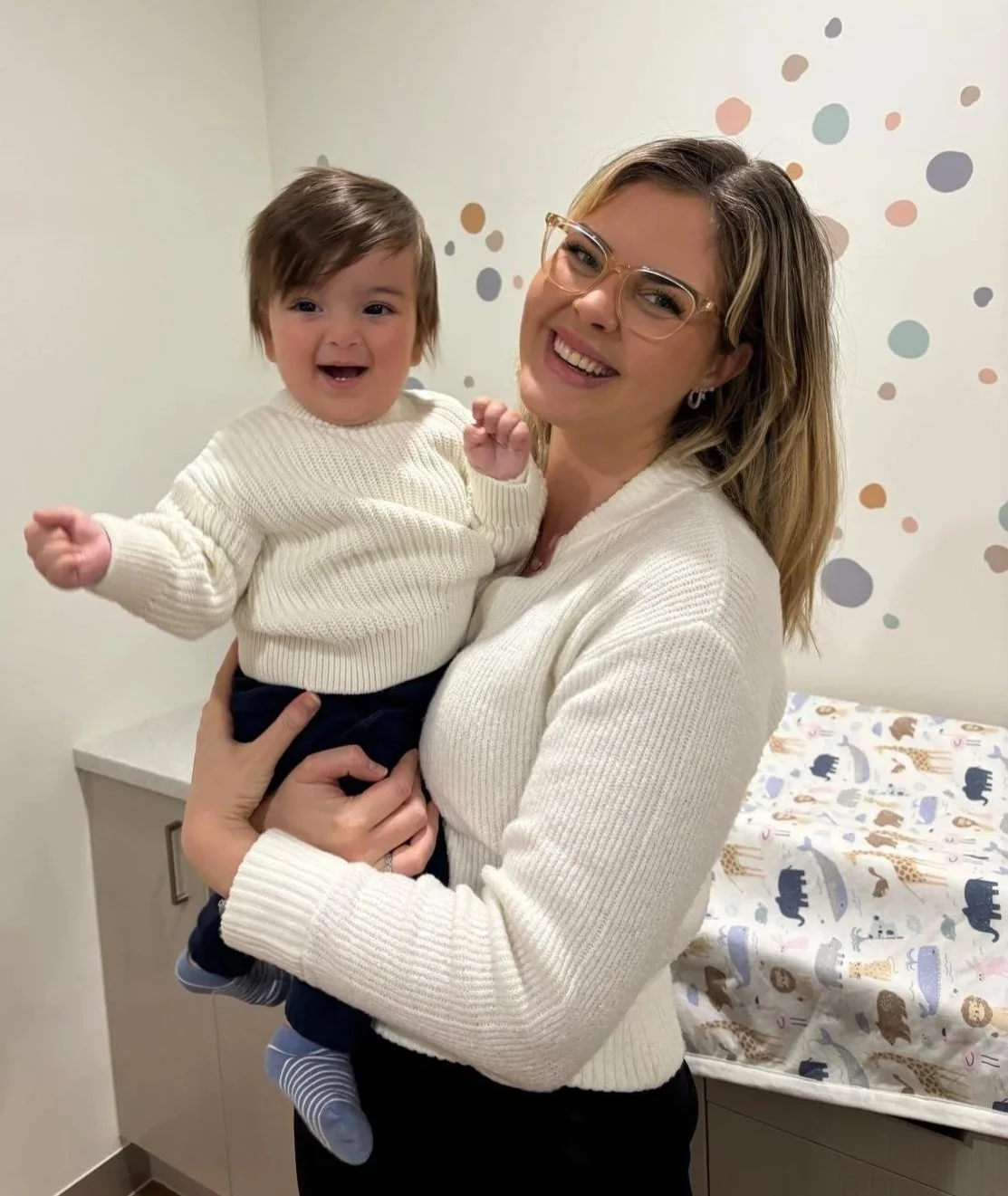 A woman with glasses holding a smiling toddler in a cream sweater, with a changing table and animal-themed mattress in the background.
