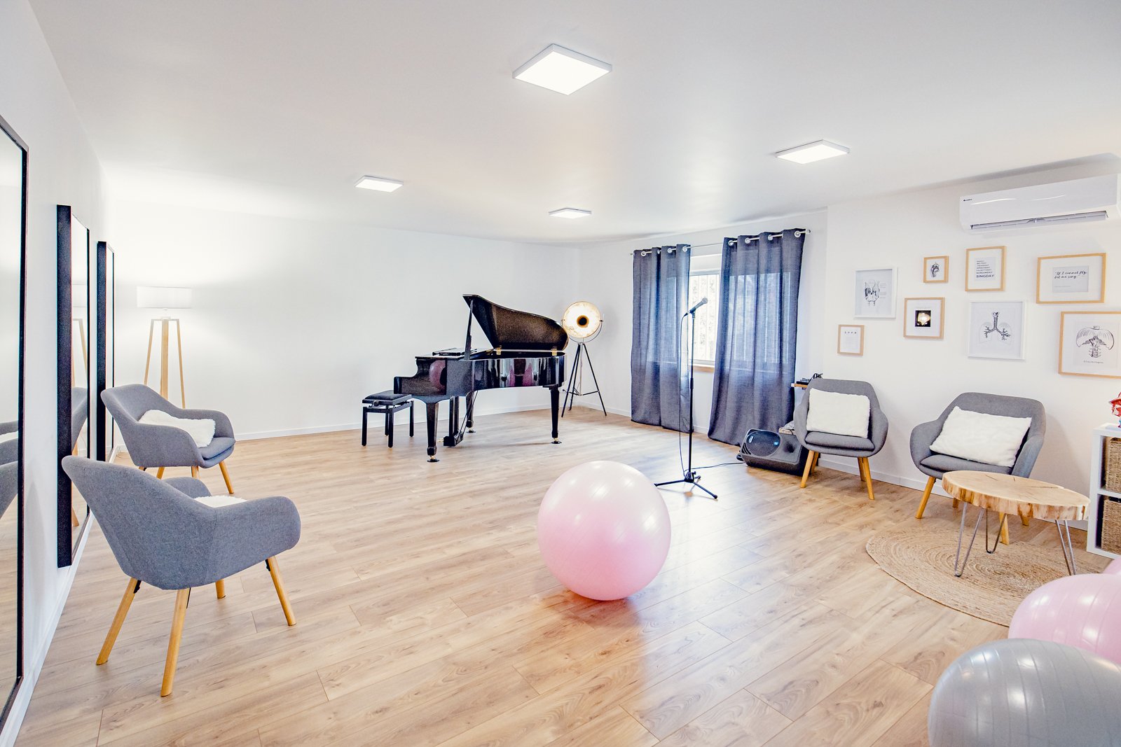 Salle de chant avec un piano à queue noir, fauteuils gris, lampadaire, mur décoré avec cadres, ballons de fête, rideaux et microphone.