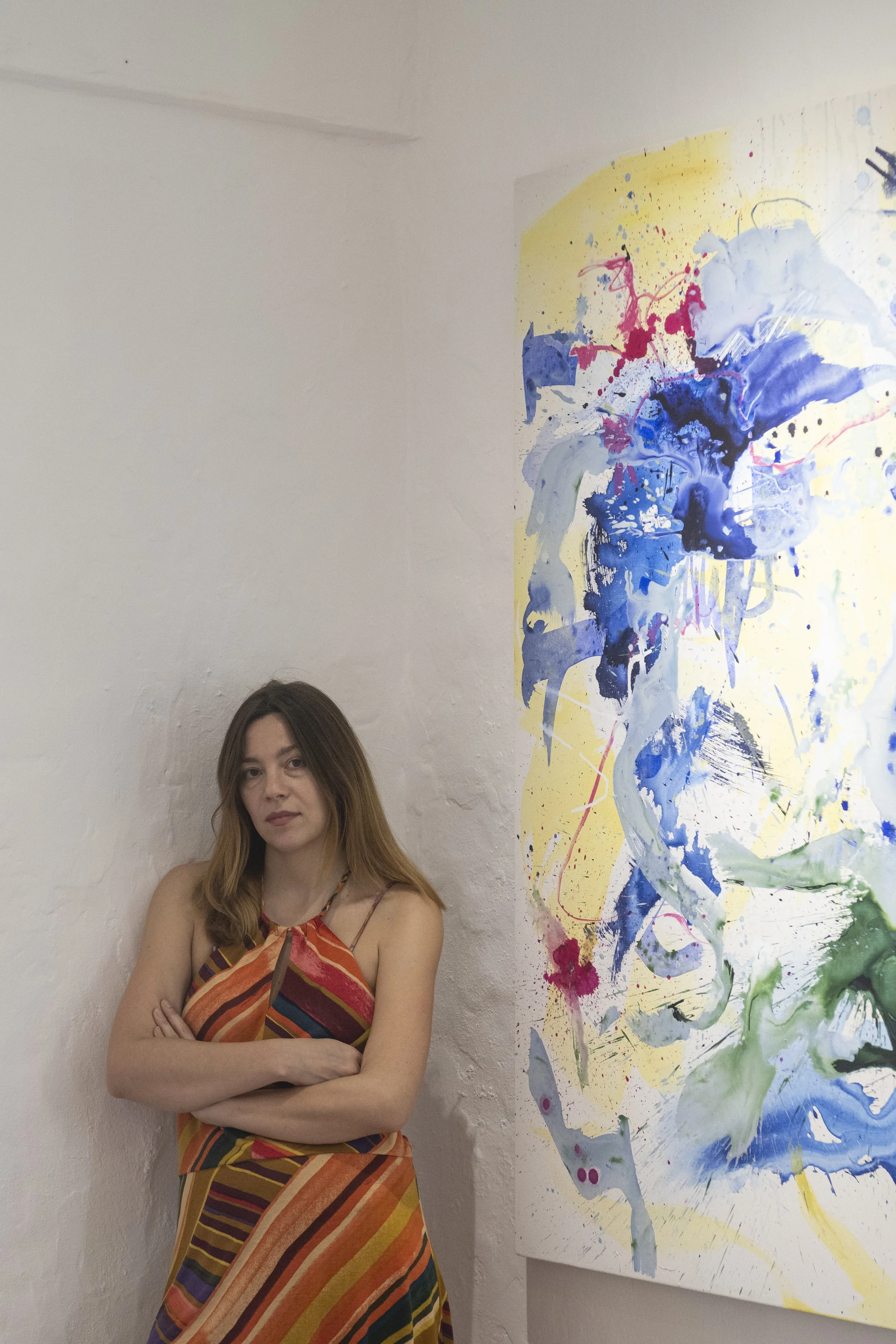A woman stands with arms crossed next to an abstract colorful painting on a white wall.