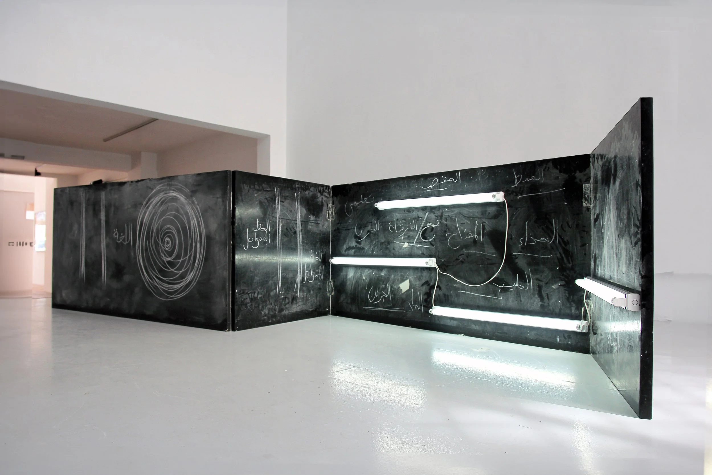 Black chalkboard with white Arabic writing and concentric circle patterns, illuminated by fluorescent lights, in an art gallery setting.