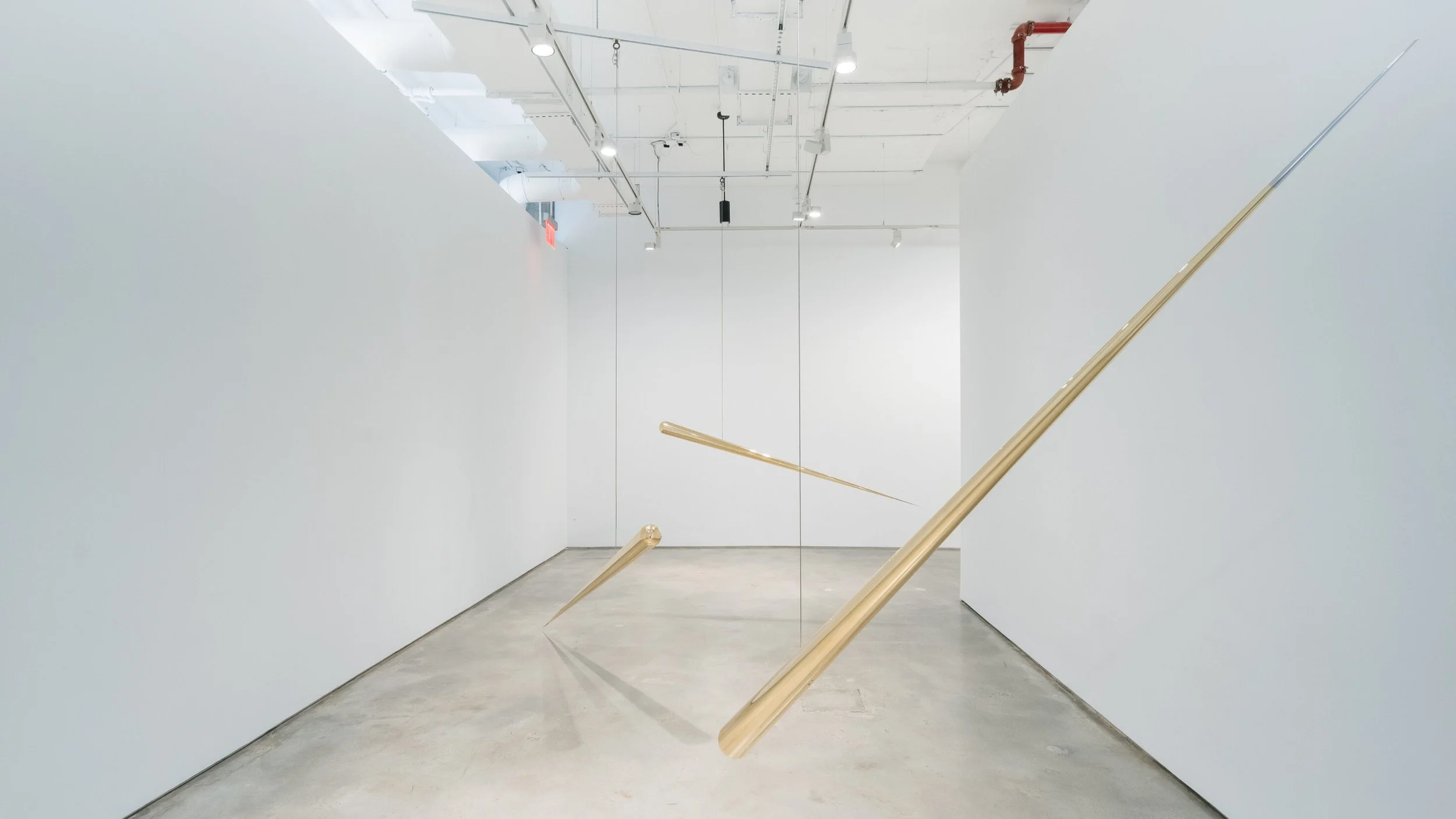Minimalist art installation featuring three elongated, gold-colored objects suspended vertically in a white gallery space with a plain white wall and ceiling with visible pipes and lighting.