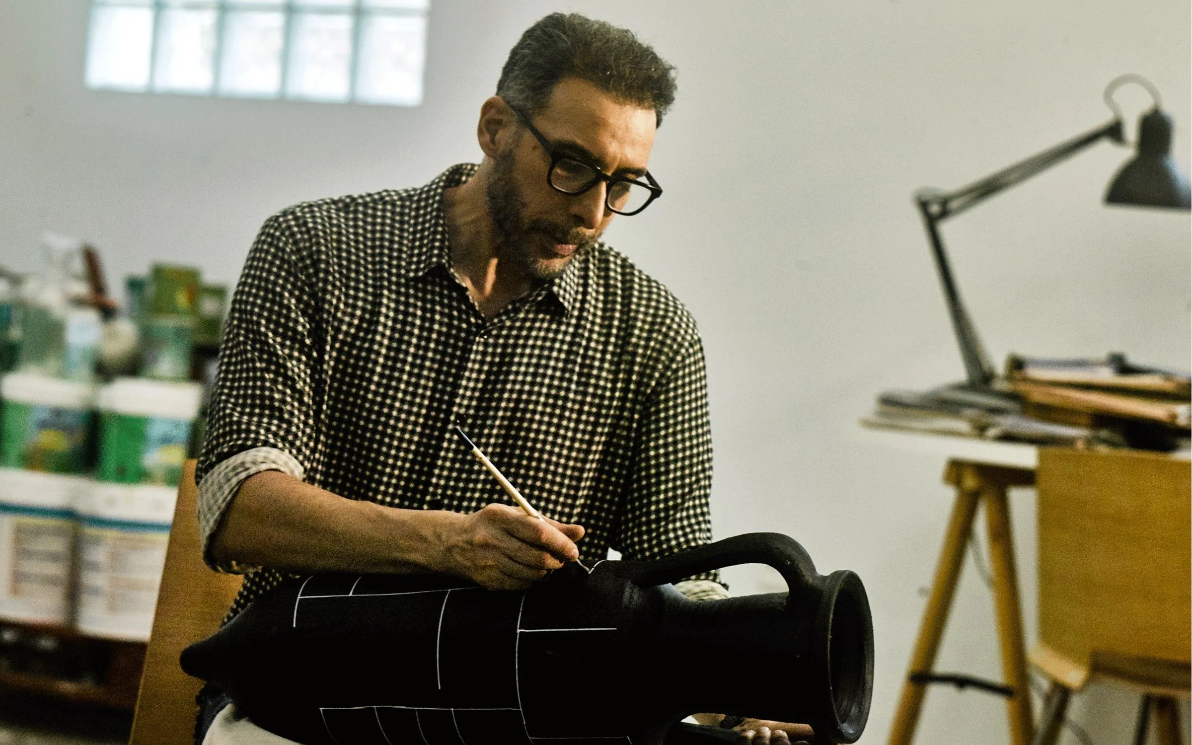 Man working on a sculpture in a workshop, with art supplies and furniture in the background.