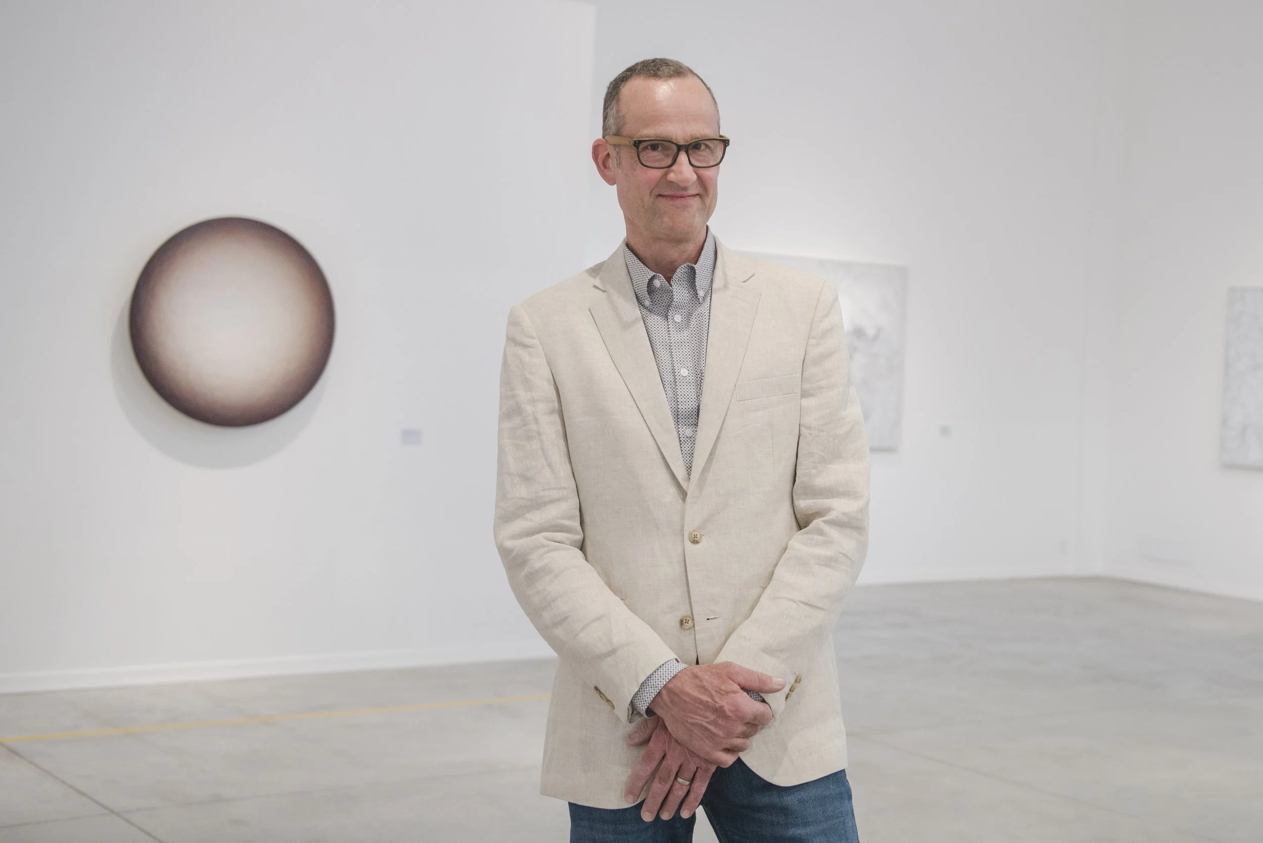 A man with glasses and short hair, wearing a beige blazer and patterned shirt, standing with hands clasped in an art gallery. Abstract circular paintings are on the white walls behind him.