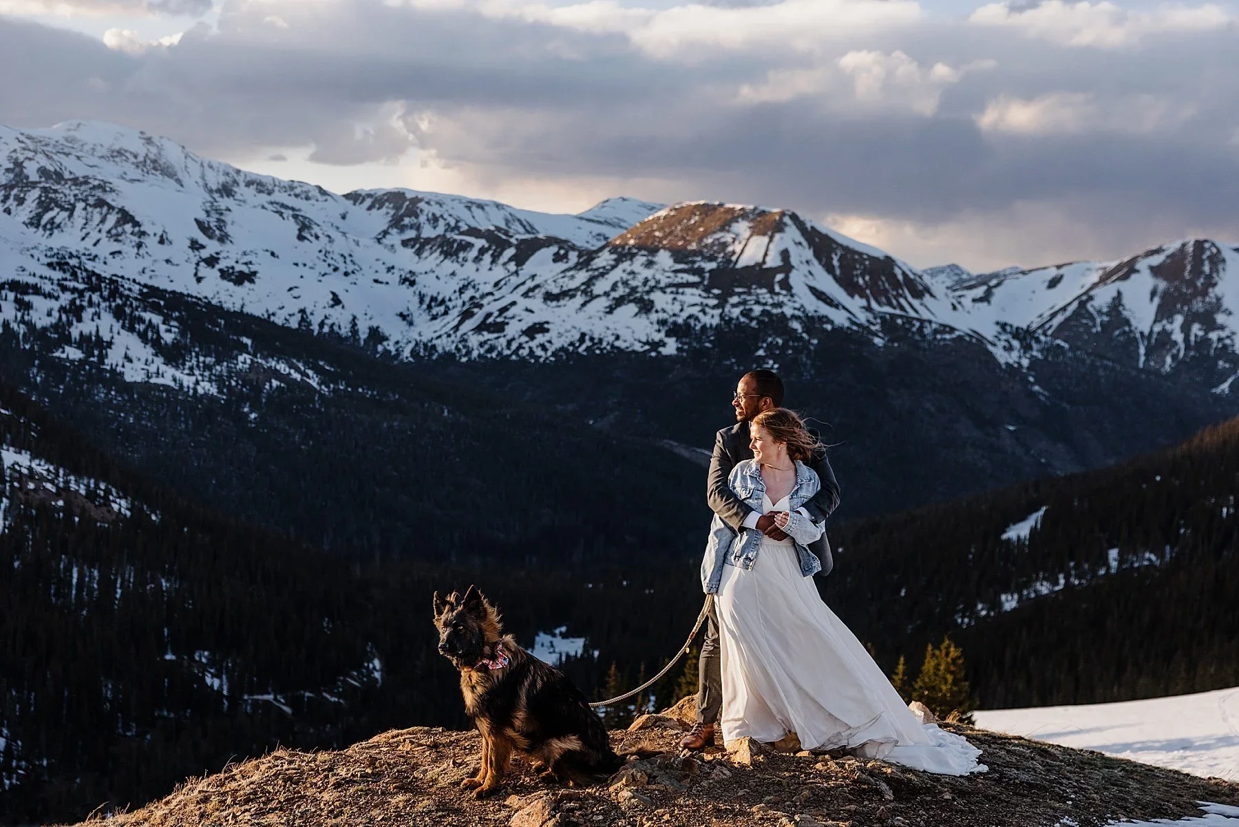 A couple stands with their dog on a snowy mountaintop