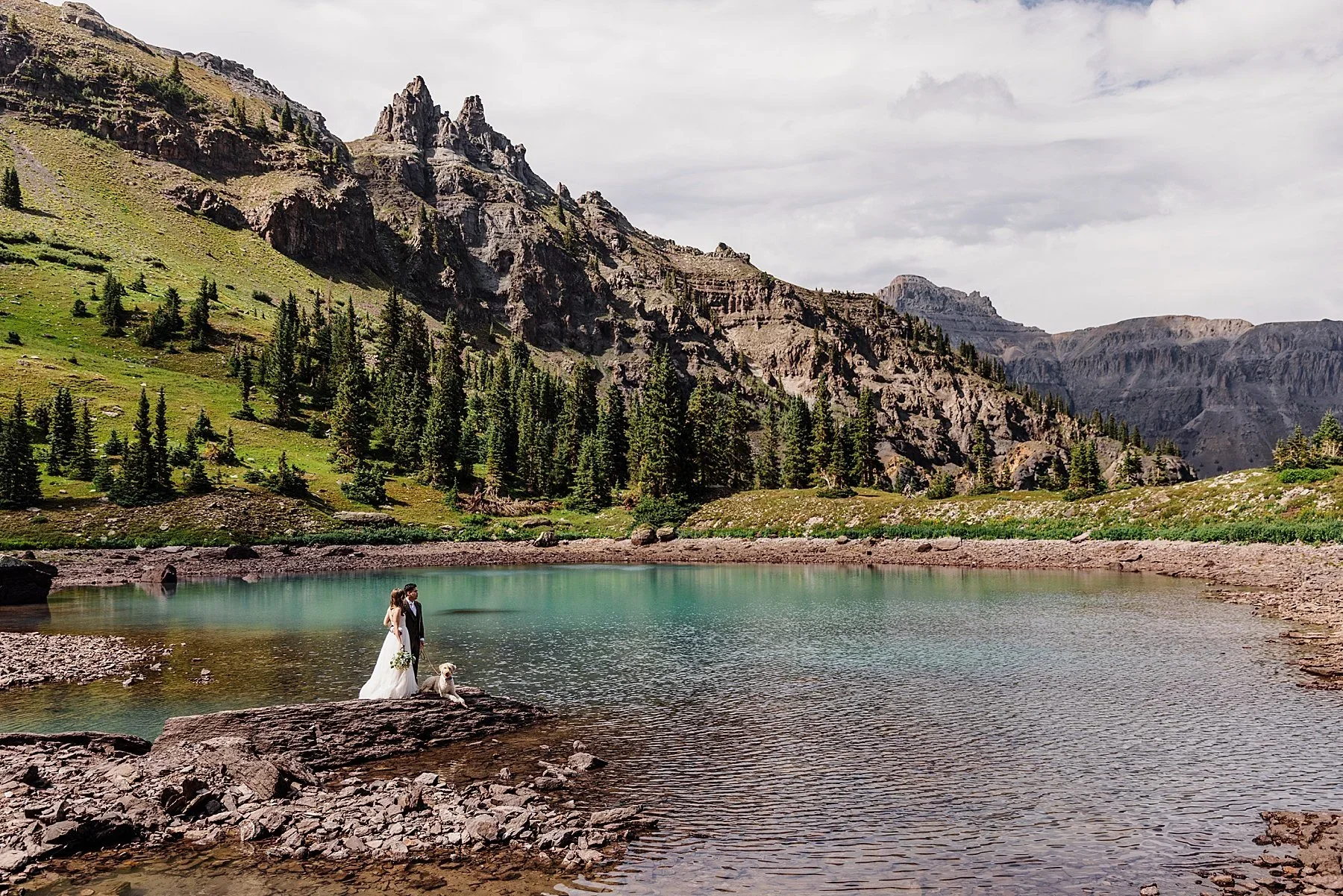 Kelsey and Han sit on a picnic blanket in front of a blue alpine lake, surrounded by mountains. Their dog lays next to them and looks back at the camera.