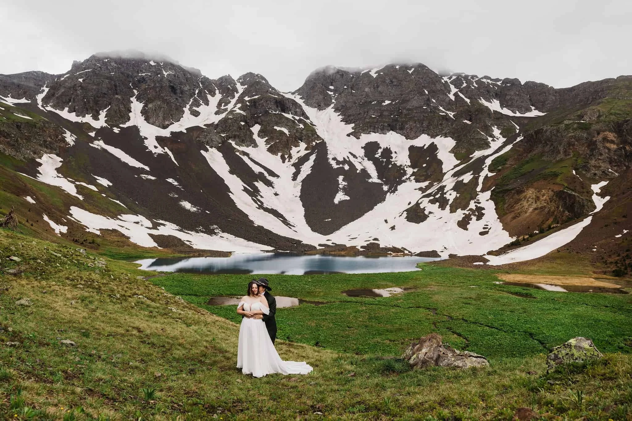 A couple cuddles together in front of an alpine lake