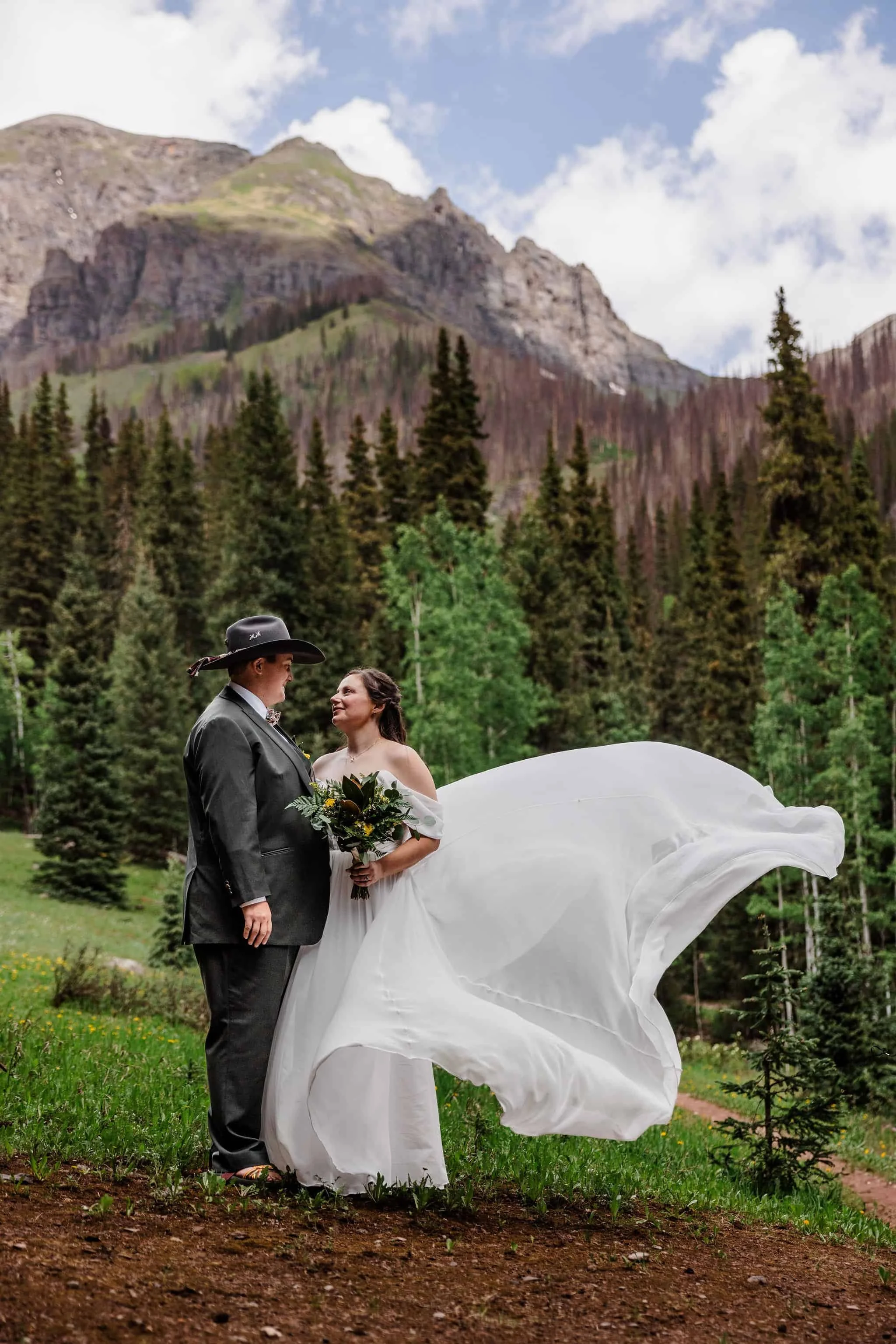 A couple stands together, the bride on the right's dress flows in the wind