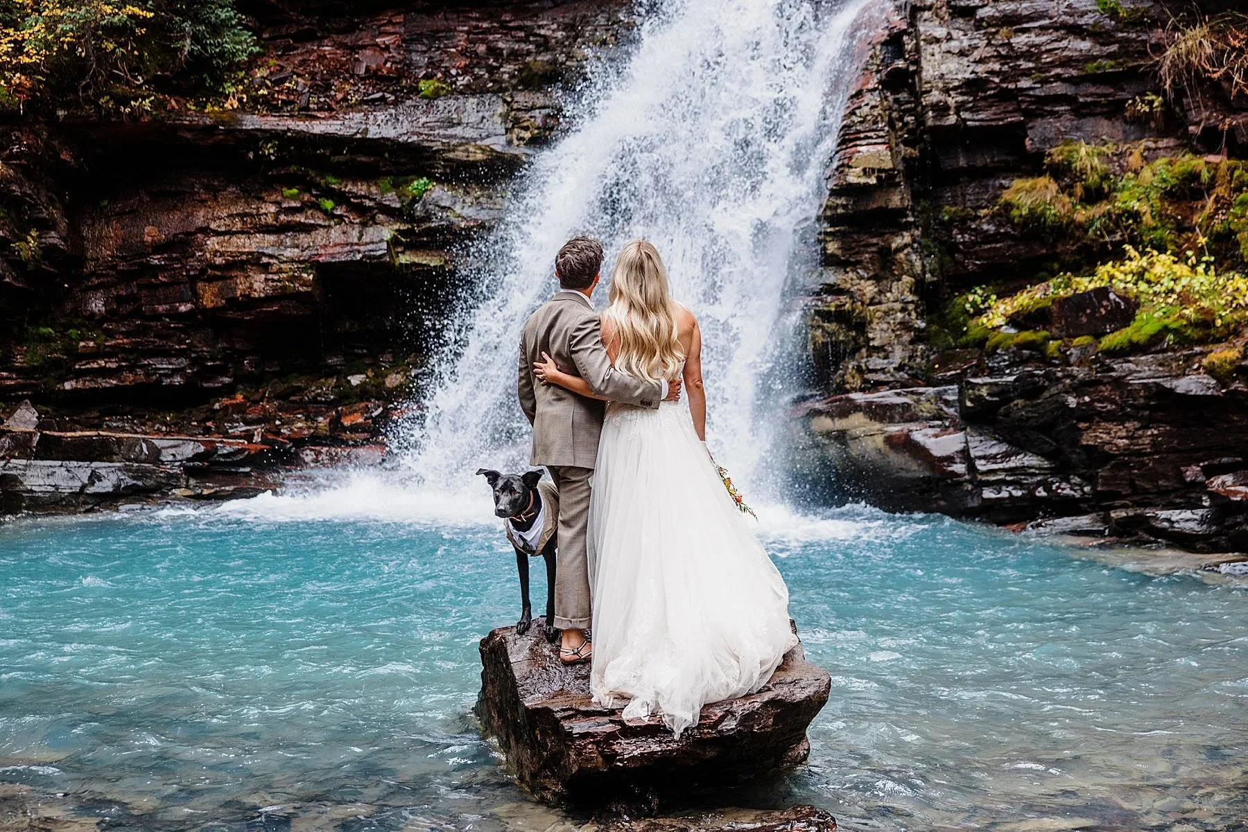 A couple stands with their dog in front of a waterfall