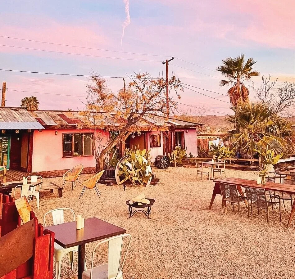 Outdoor dining area with tables, chairs, cacti, palm trees, and a pink-colored building, under a pink and purple sky at sunset.