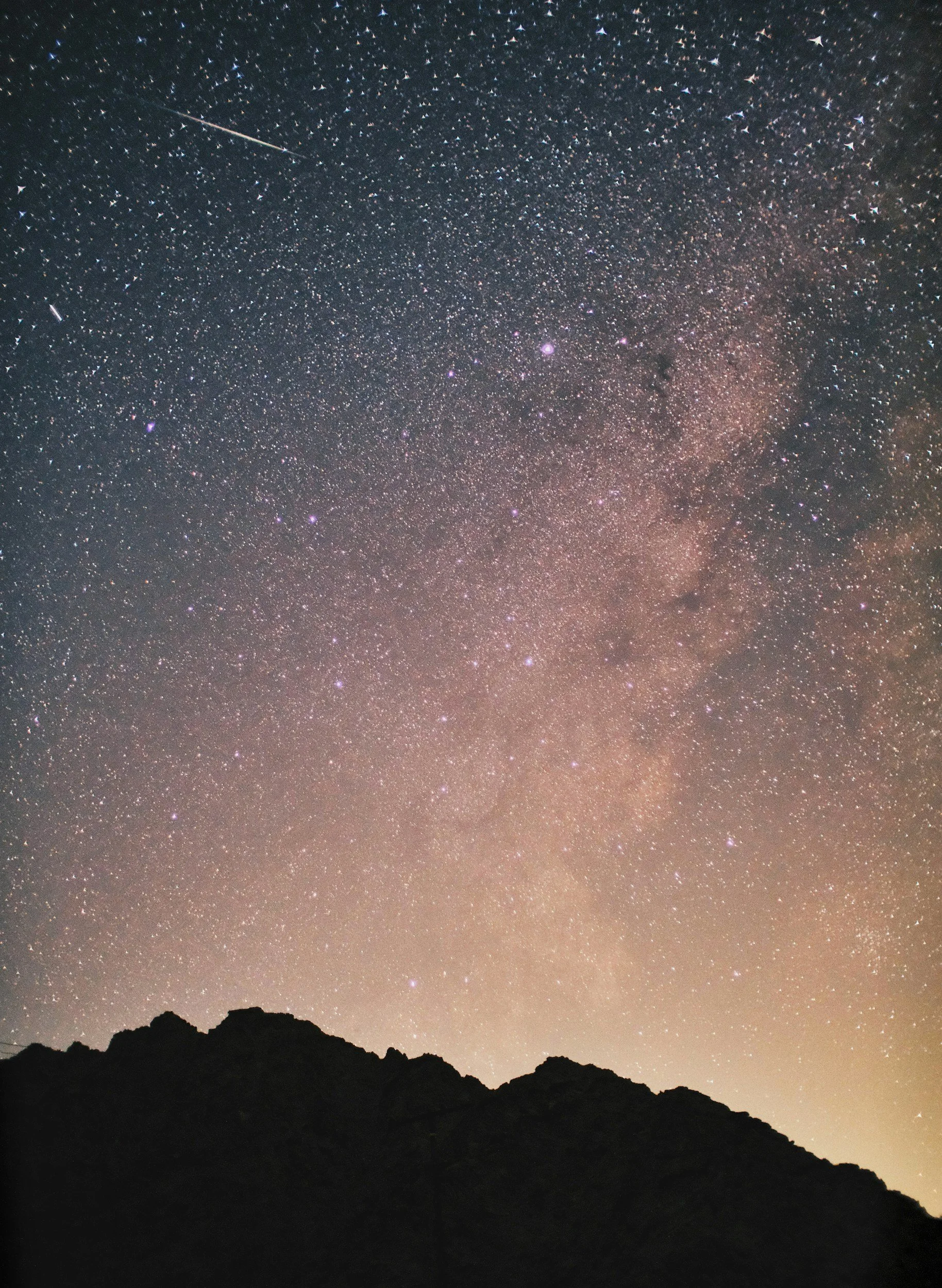 Night sky filled with stars, a shooting star, and the Milky Way galaxy above a silhouette of mountain range.