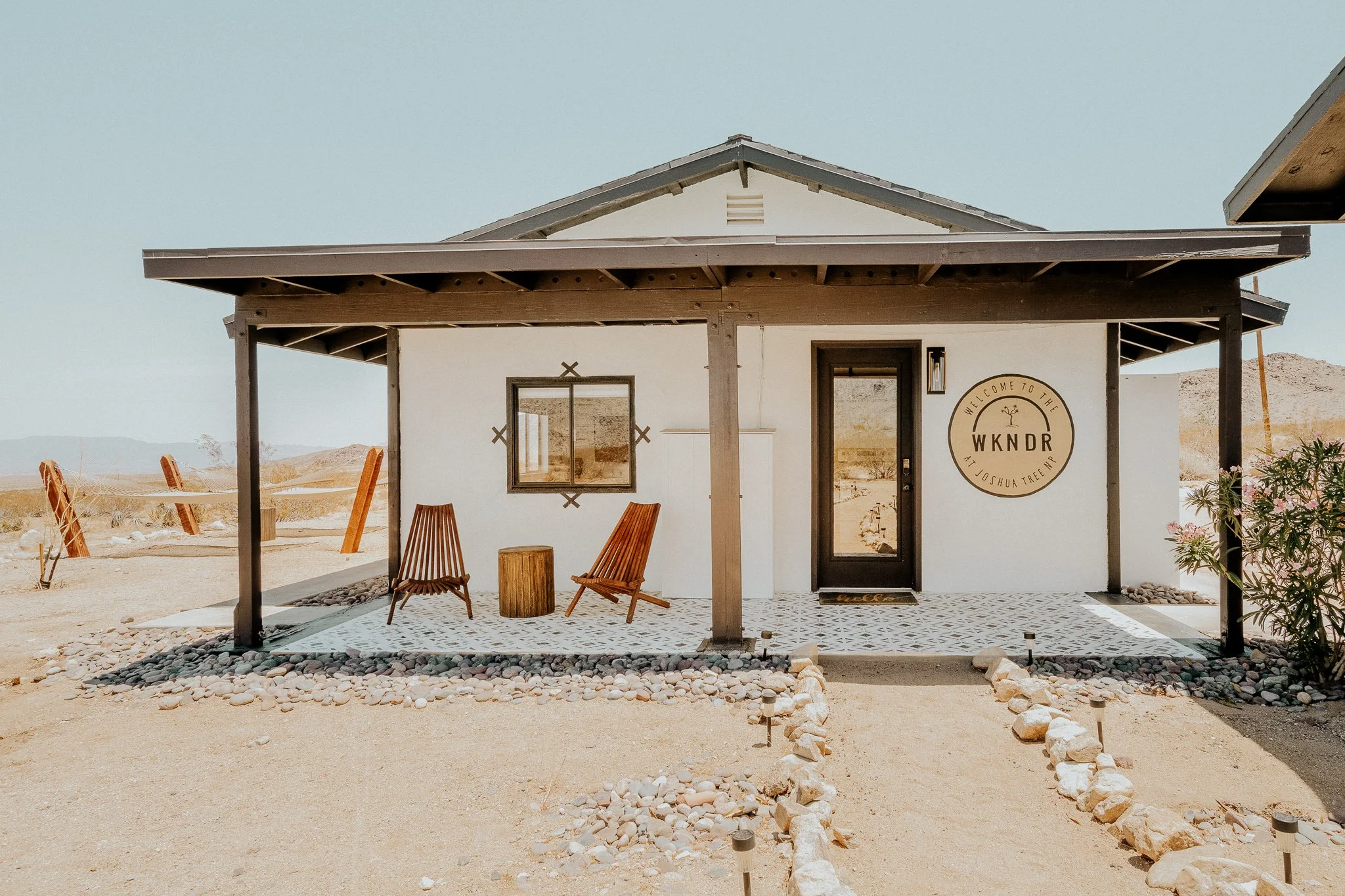 Desert home with a covered porch, two wooden chairs, a tree stump table, and a sign that says "Welcome to the WKNDR" near Joshua Tree, California.