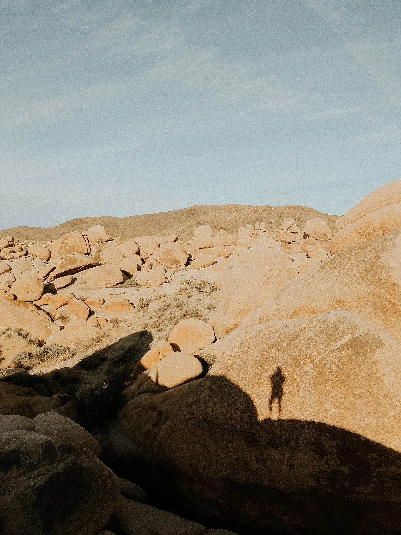 Desert landscape with large rocks and boulders, with a person's shadow cast on a rock, under a blue sky with clouds.