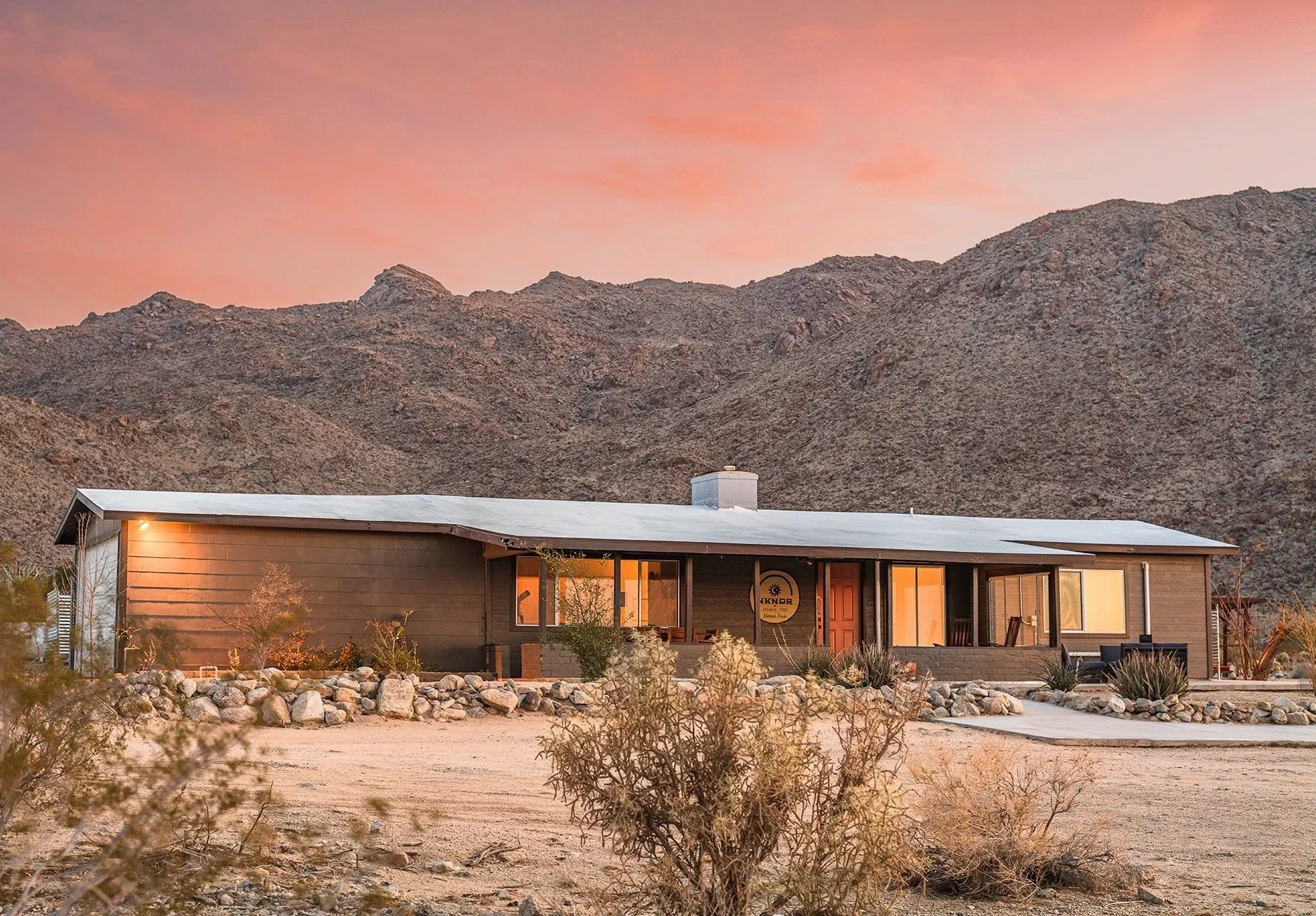 A single-story house in a desert landscape during sunset, with mountains in the background and rocks along the front yard.
