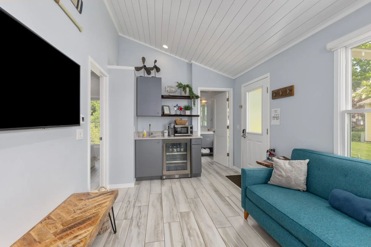 Interior of a cozy living space with a blue sofa, a mounted large flat-screen TV, a small kitchenette with a mini fridge, shelves, and a microwave, and a door leading outside, with large windows letting in natural light.