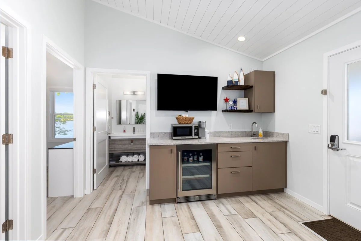 A small kitchenette with a microwave, mini fridge, sink, open shelves, and a mounted TV on white walls in a bright room.