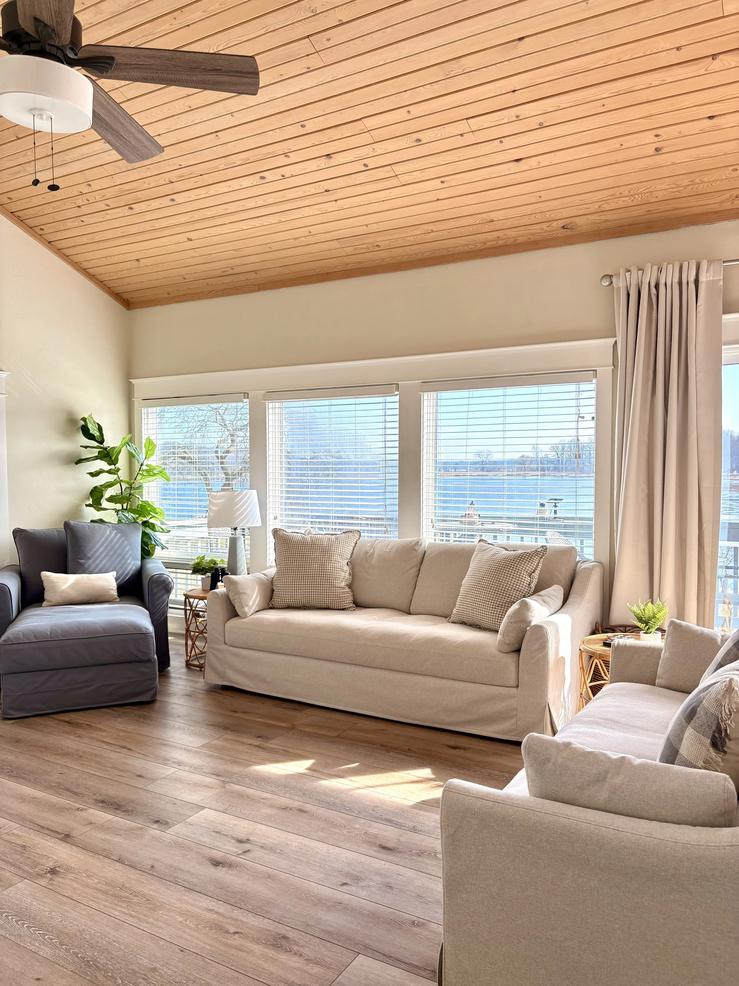 A living room with three large windows, beige and gray sofas, a plant, a wooden side table, a white table lamp, a ceiling fan with wooden blades, and light-colored wood flooring.