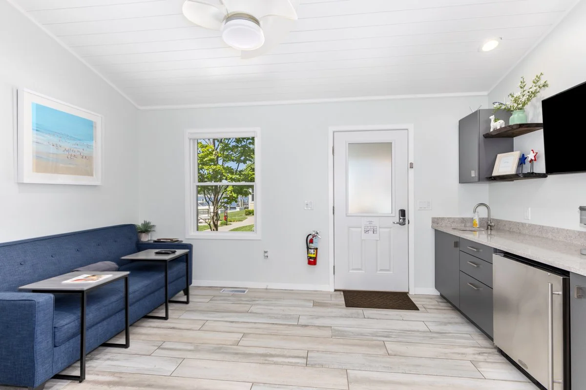 Interior of a small, modern kitchenette and seating area with light-colored wood flooring, a blue sofa, two small side tables, a white door with frosted window, window showing greenery outside, and a gray kitchen counter with built-in sink and applia