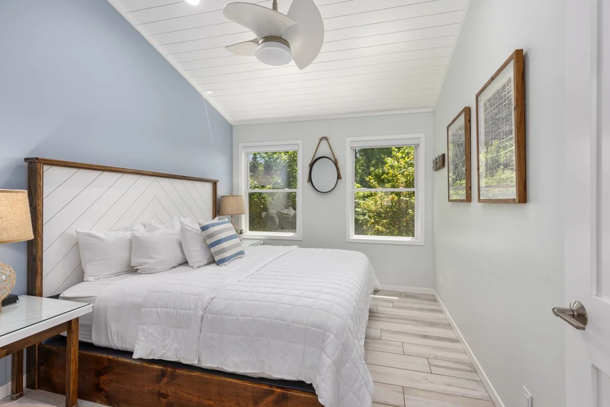 Bedroom with a white bed, wooden headboard, two windows, and artwork on the walls, illuminated by natural light.